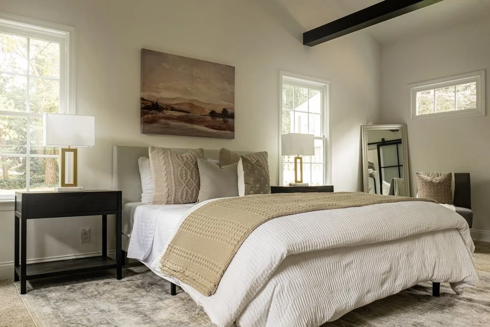 Master bedroom full view with neutral beige throw pillows and blankets anchored with two black nightstands and large windows.