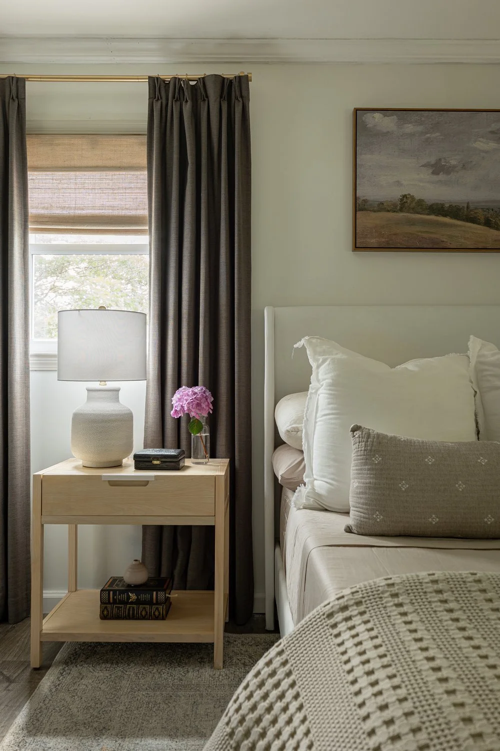 Neutral, modern bedroom details of half white upholstered bed, wooden nightstand, and layered window treatments