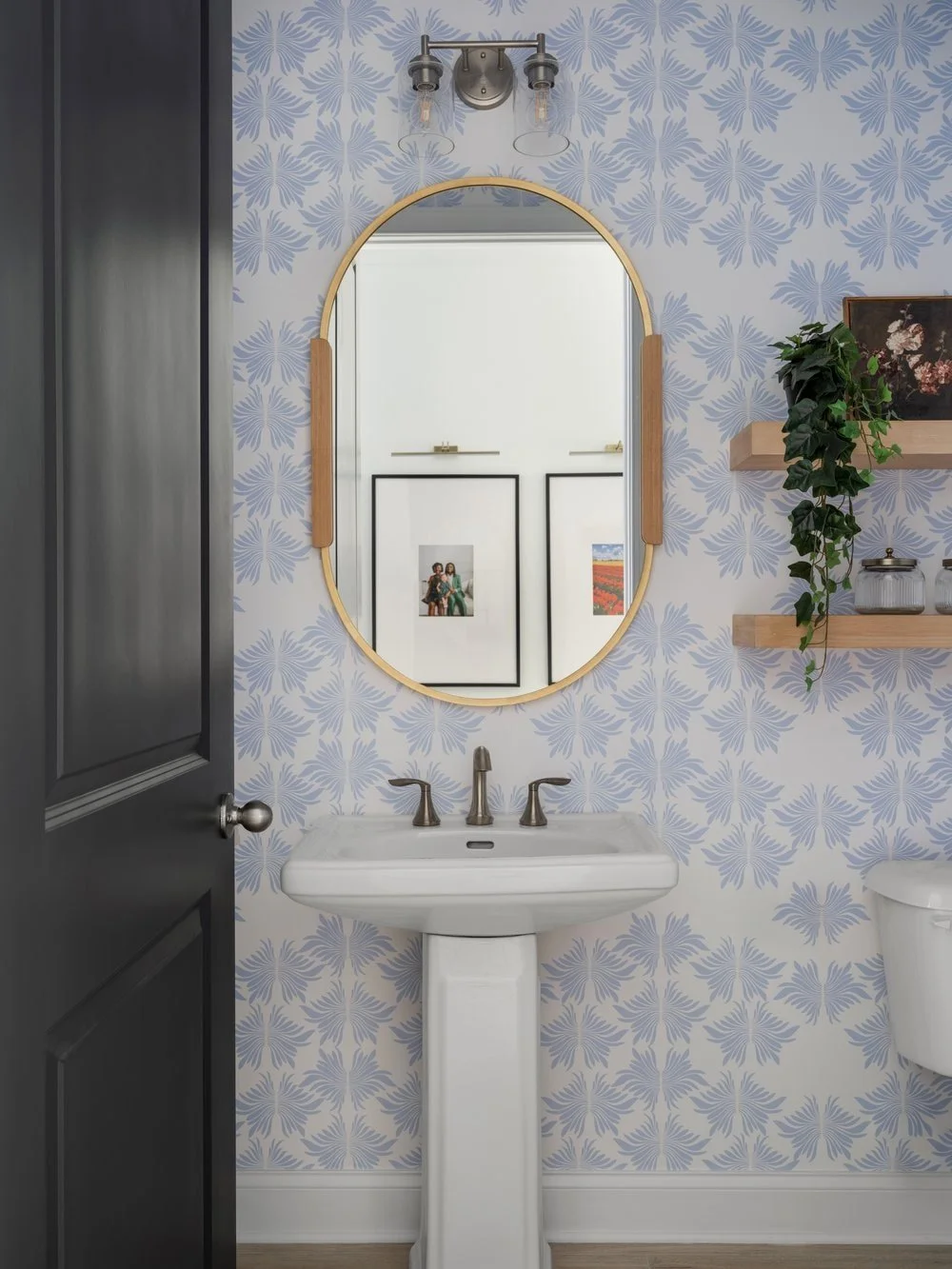 Powder bathroom in Georgia family home with coastal blue wallpaper and an oval mirror reflecting the hallway frames