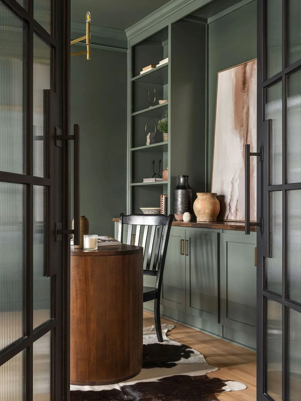 Custom steel doors detailed with fluted glass framing dark green color drenched home office and walnut rounded office desk with cow hide rug