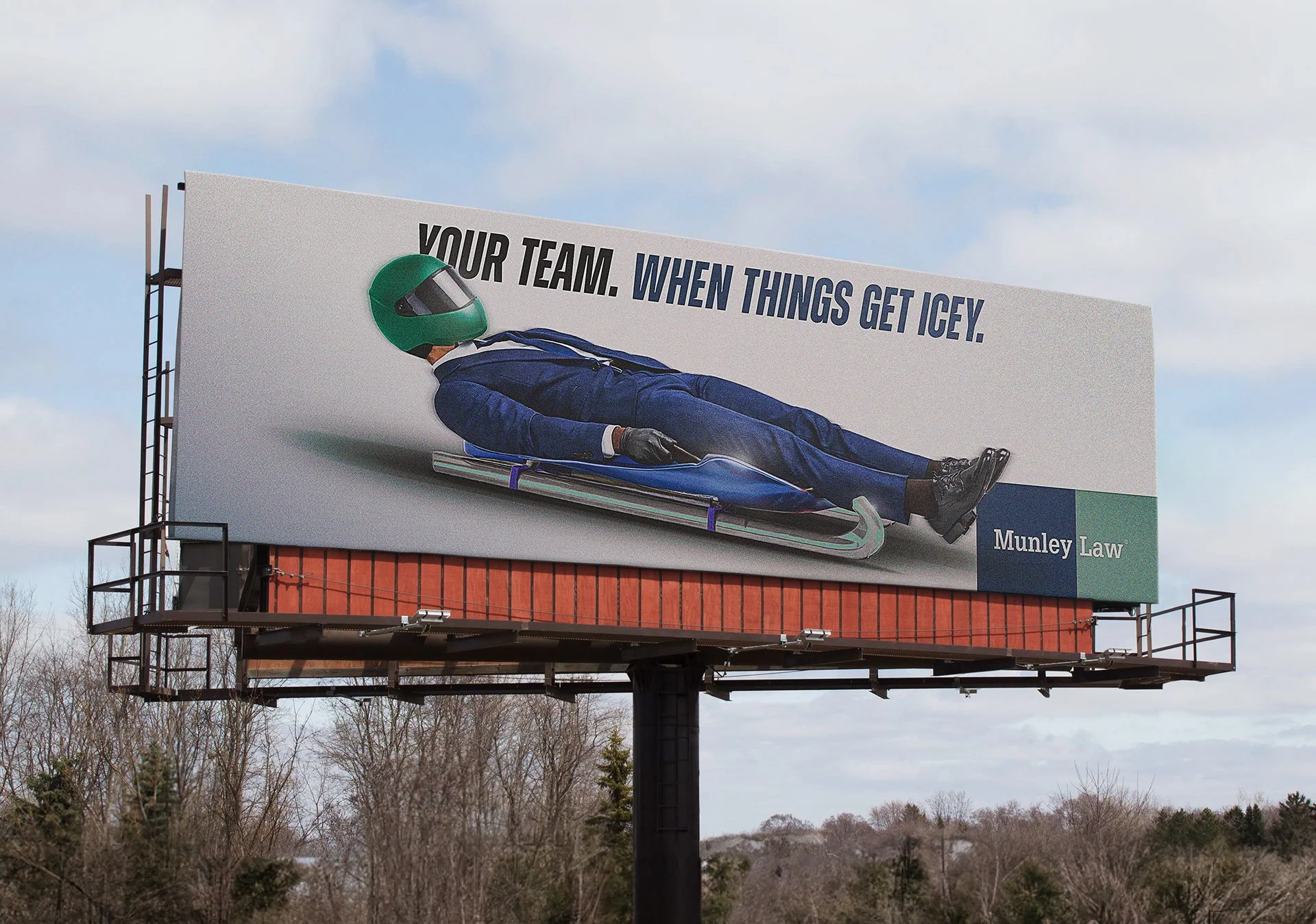 Billboard advertising a law firm with a man in a suit lying on a sled wearing a green helmet and goggles, with the text "Your team. When things get icey."