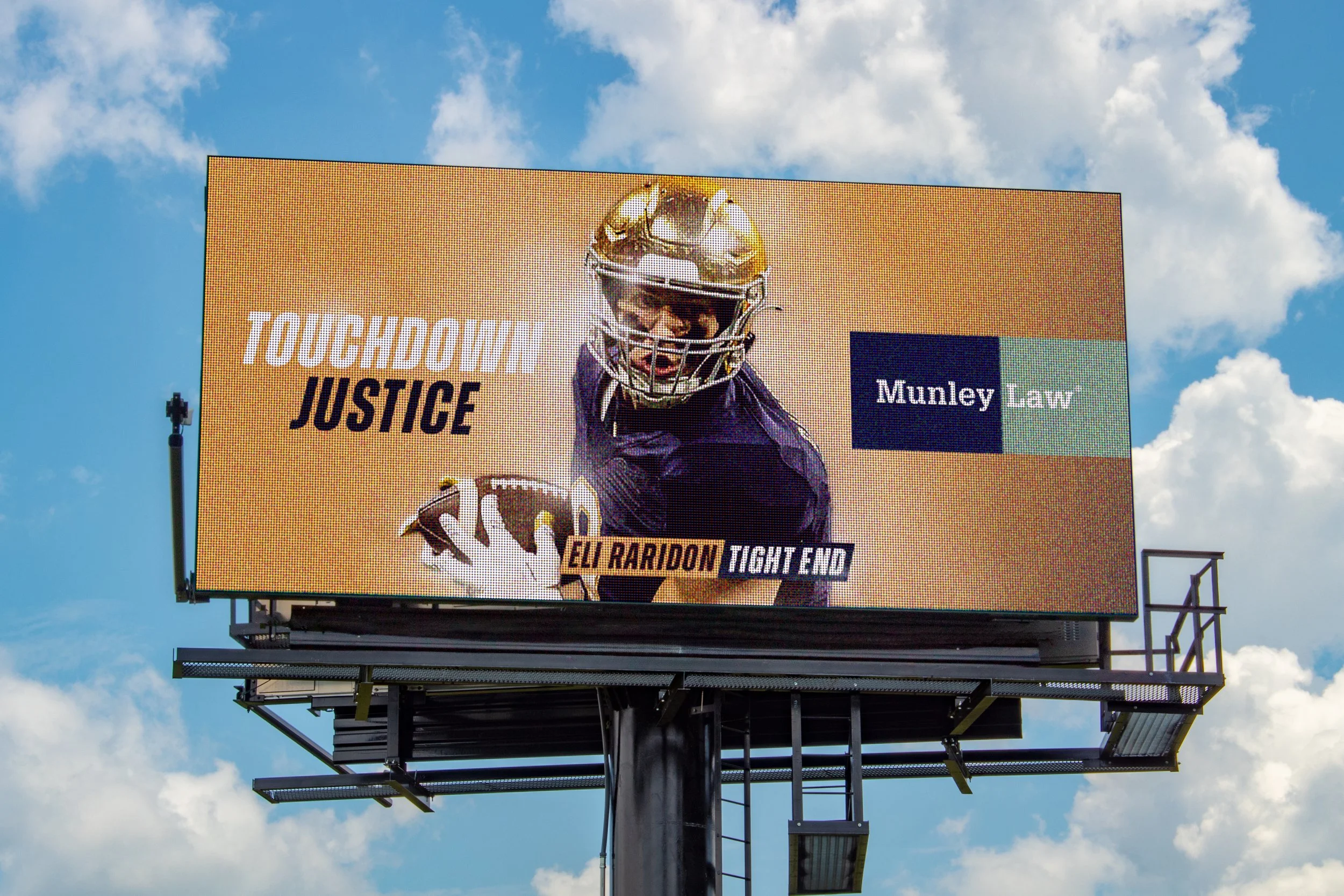 Large outdoor digital billboard celebrating a football player's touchdown, featuring a photo of Elg Pipe Raridon in a football helmet and uniform, with the words 'Touchdown Justice' and 'Munley Law' displayed.