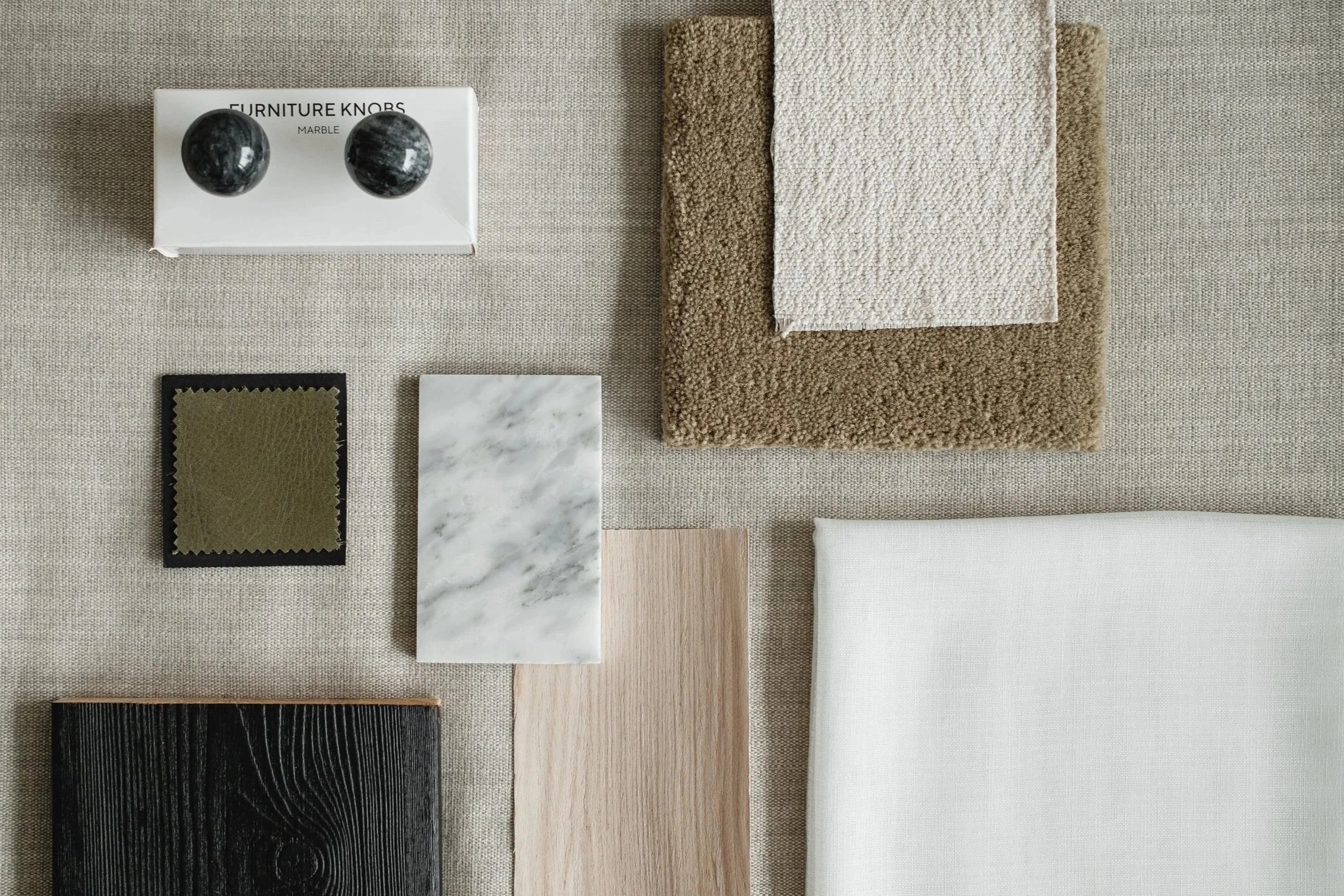 A collection of interior design materials including black marble furniture knobs, fabric swatches, carpet, various wood and marble samples, and textured fabrics laid out on a beige fabric background.