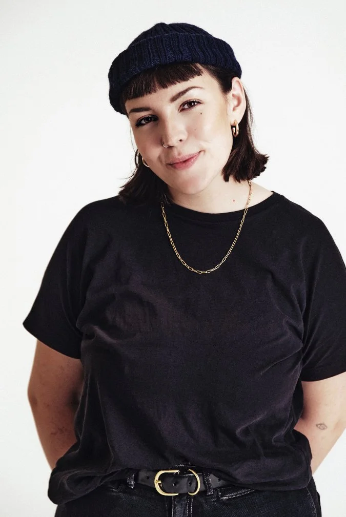 Junge Frau trägt schwarzen T-Shirt, schwarzen Hut, und hat kurze braune Haare. Tourmanagement, Eventproduktion Artist Care aus Hamburg.