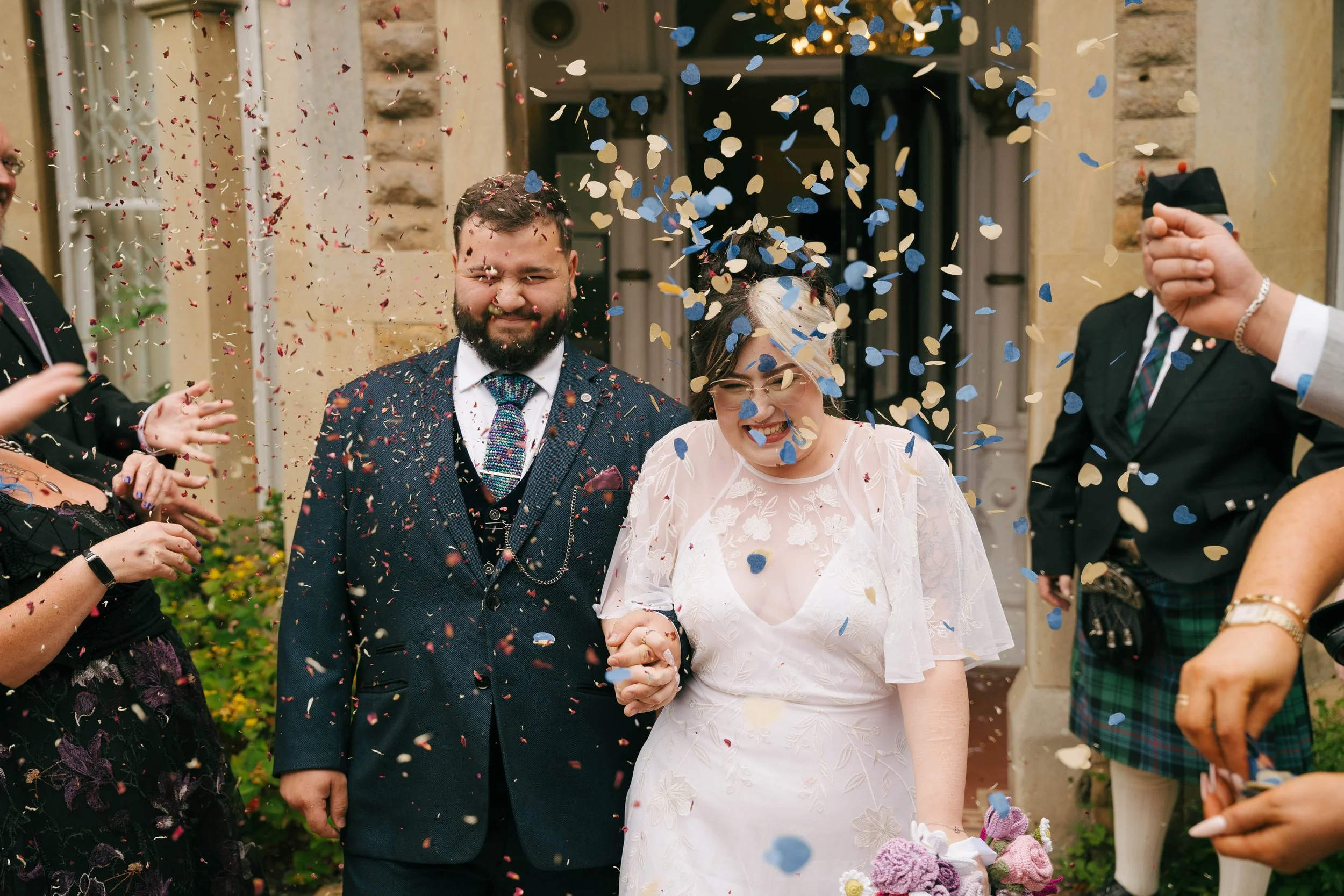 A newlywed couple is celebrated as guests throw colorful confetti during their wedding. The groom has a beard and is dressed in a dark suit with a colorful tie, while the bride wears glasses and a white lace dress. The scene is outside a building wit