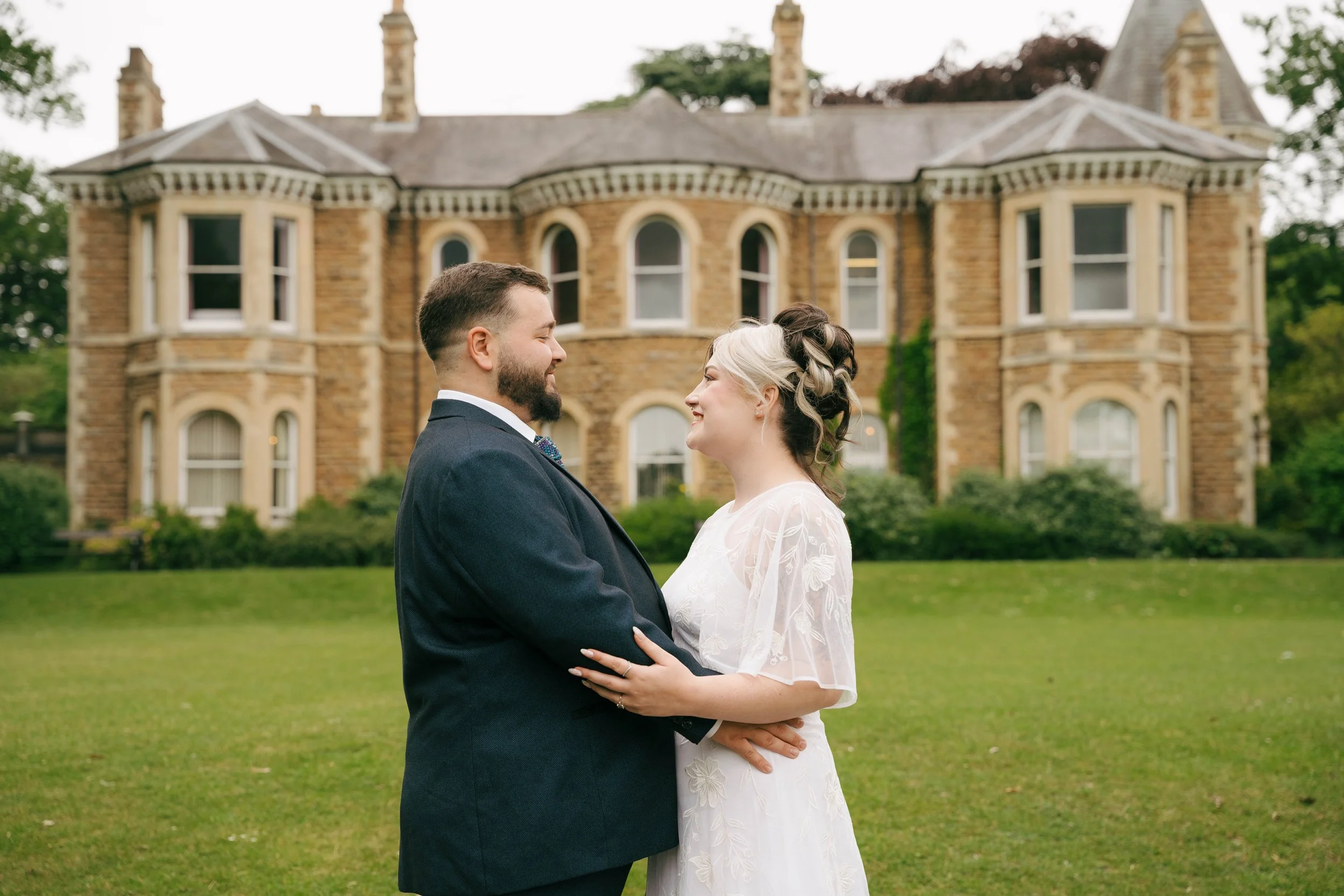 A newlywed couple smiling and holding each other in front of a large historic brick house with trees in the background.