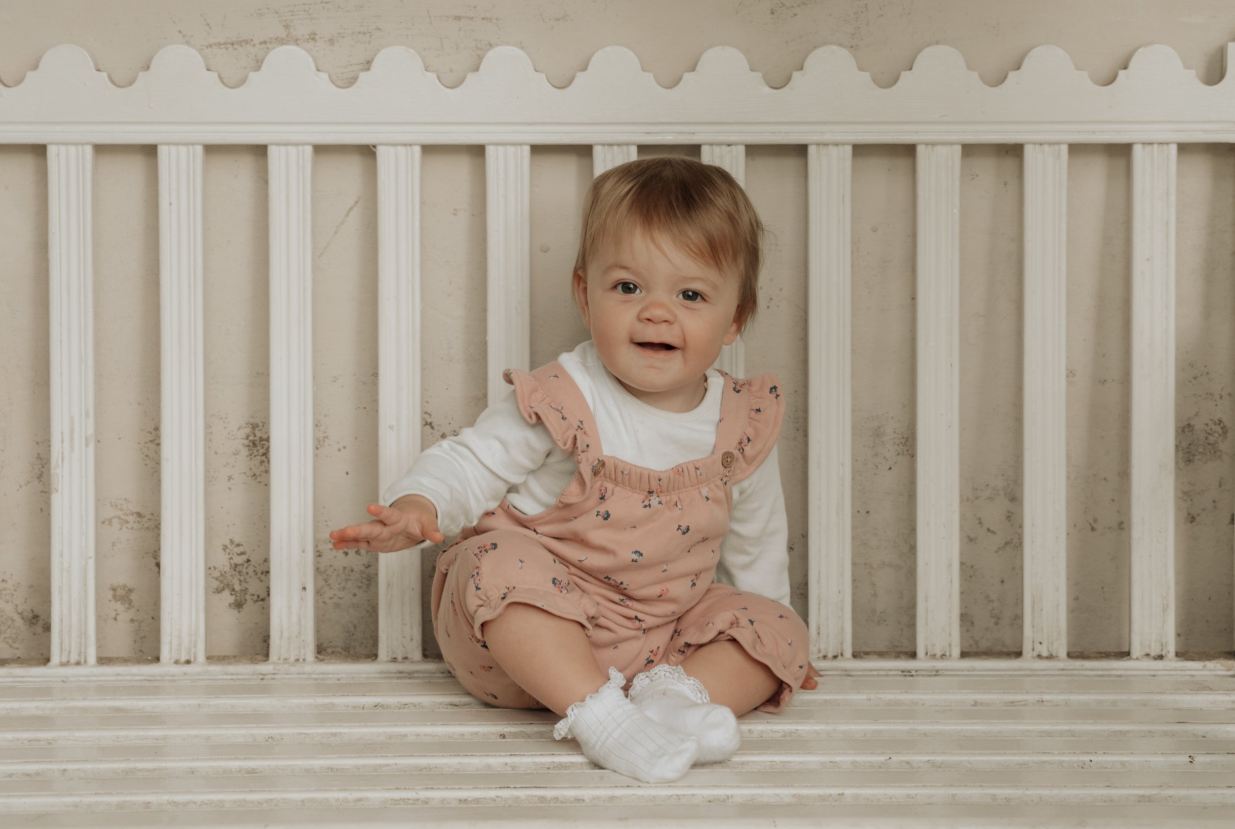 A young child with light brown hair and a white shirt, wearing a pink jumpsuit with floral print, sitting on a white wooden bench or bed with vertical slats behind and beneath them, and a beige wall with a curved border above.