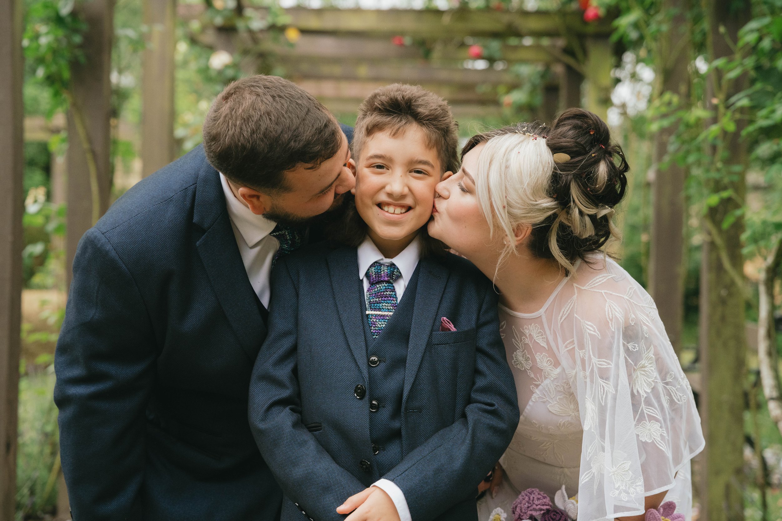 A young boy in a suit between two adults, a man and a woman, receiving kisses on the cheeks at a garden wedding ceremony.