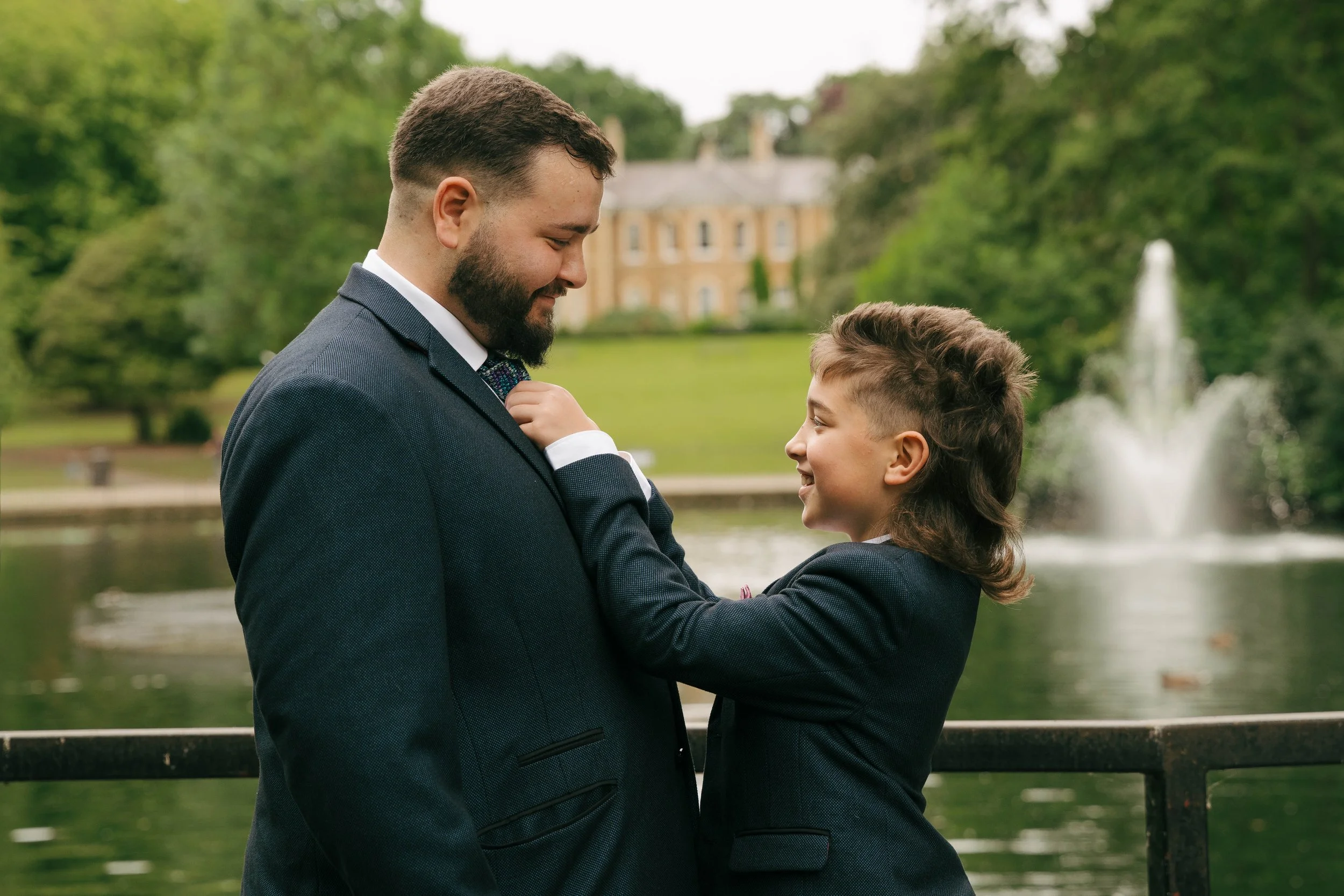 A man and a young boy, both dressed in suits, are smiling at each other outdoors near a body of water, with a large house and trees in the background.