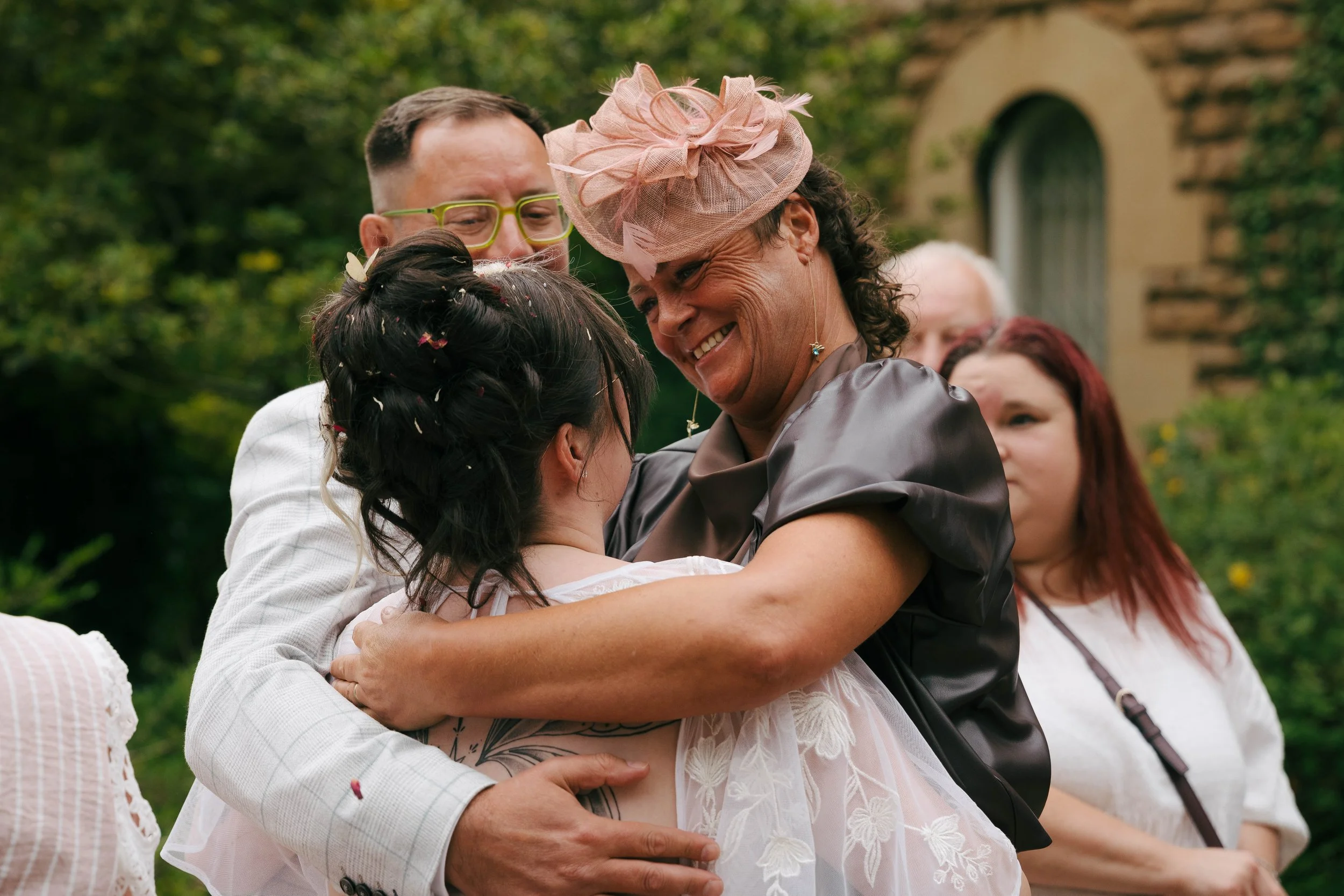 A group of people, including a woman wearing a pink fascinator hat, embracing each other outdoors during a celebration.