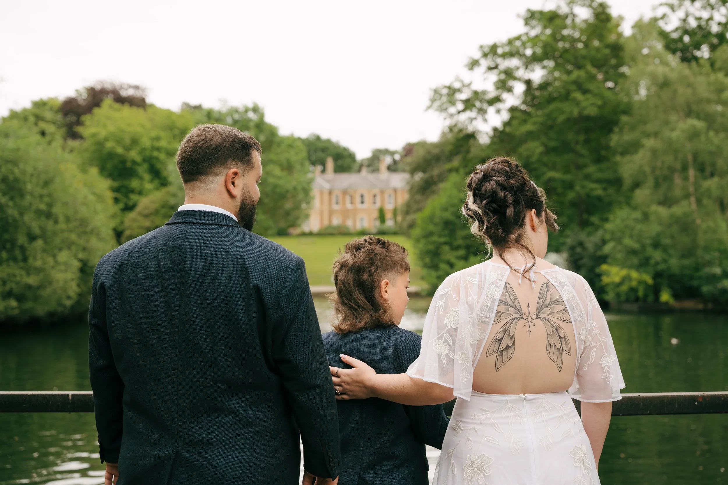 People standing by a river with a large house in the background, dressed for a wedding or special occasion.