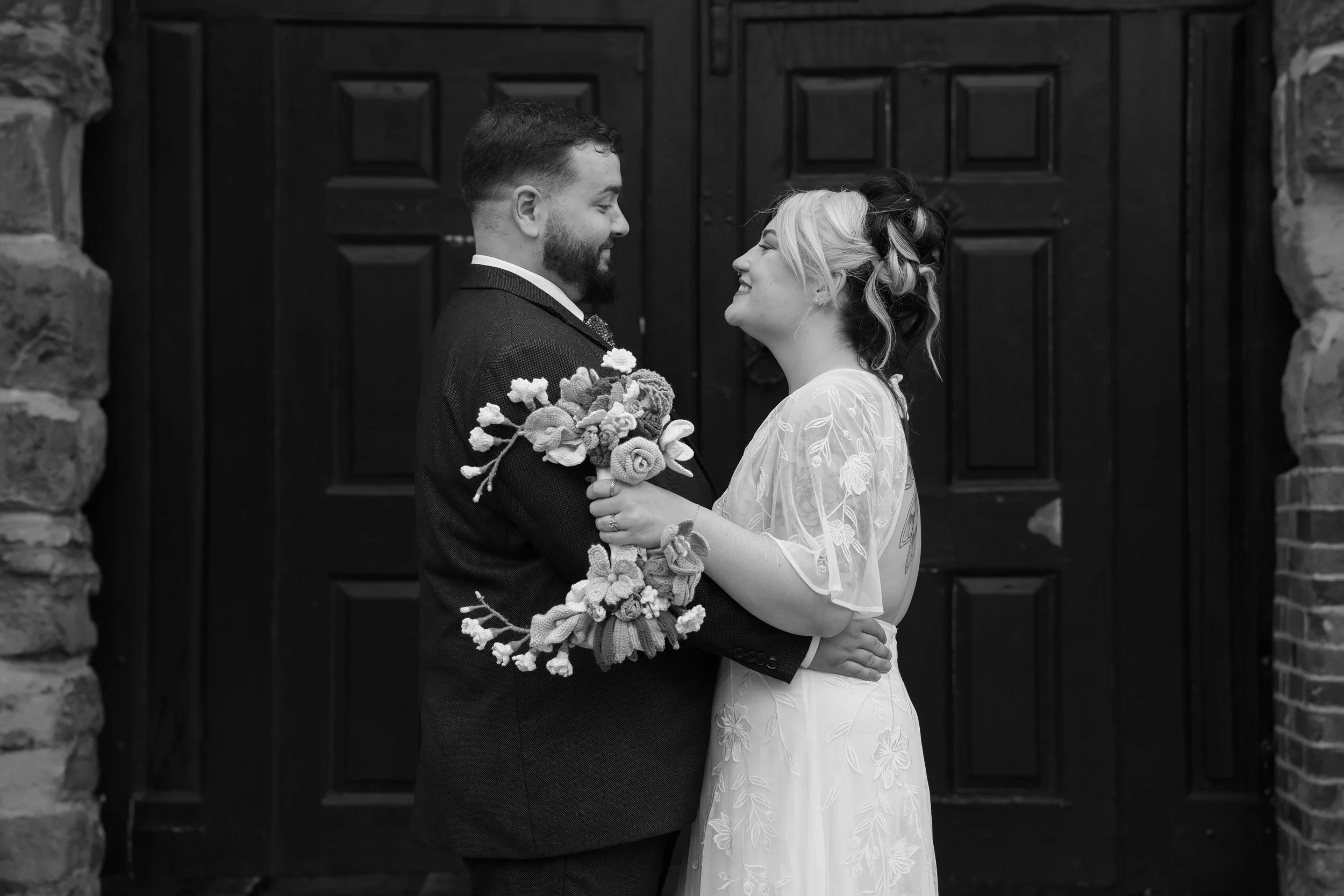 Wedding photography. A black and white photo of a bride and groom facing each other, holding hands, standing in front of a dark wooden door framed by stone walls, smiling.