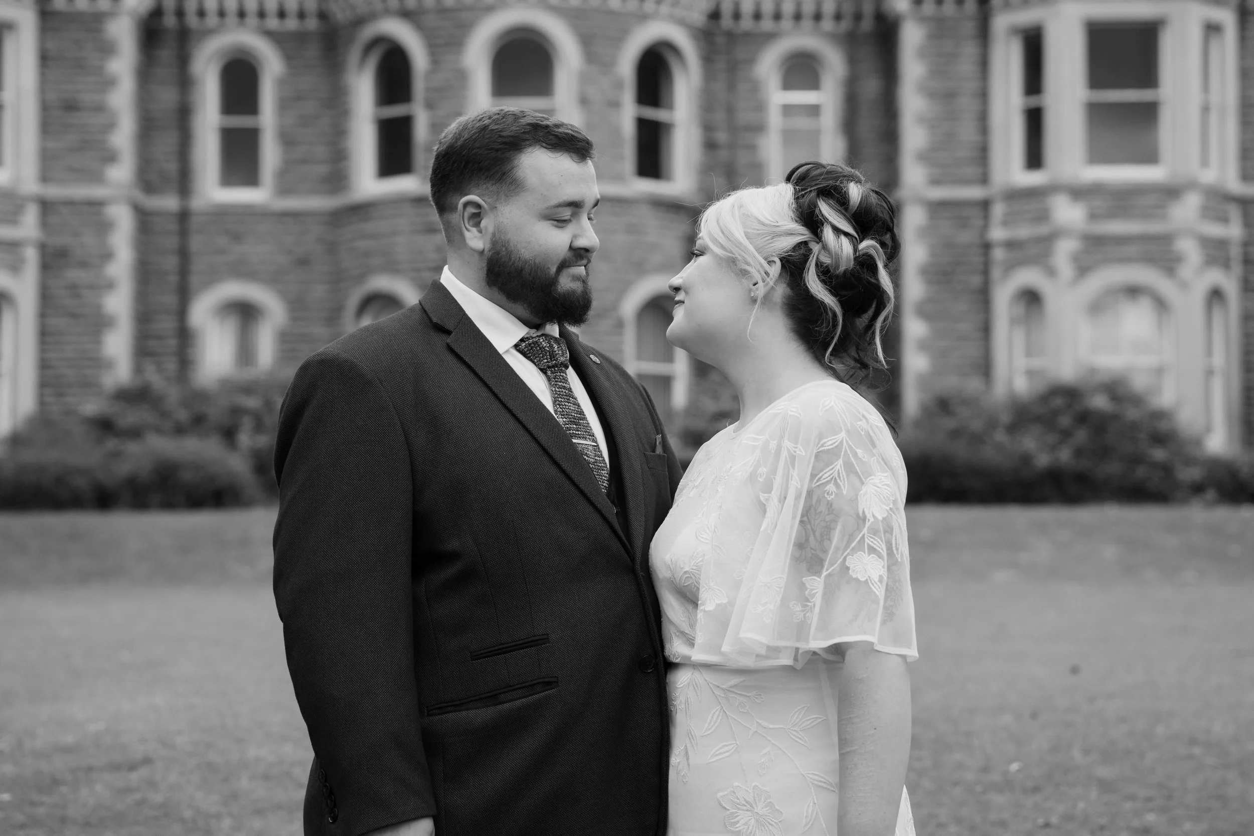 A black and white photograph of a couple standing outdoors in front of a stone building with multiple arched windows, gazing at each other lovingly.