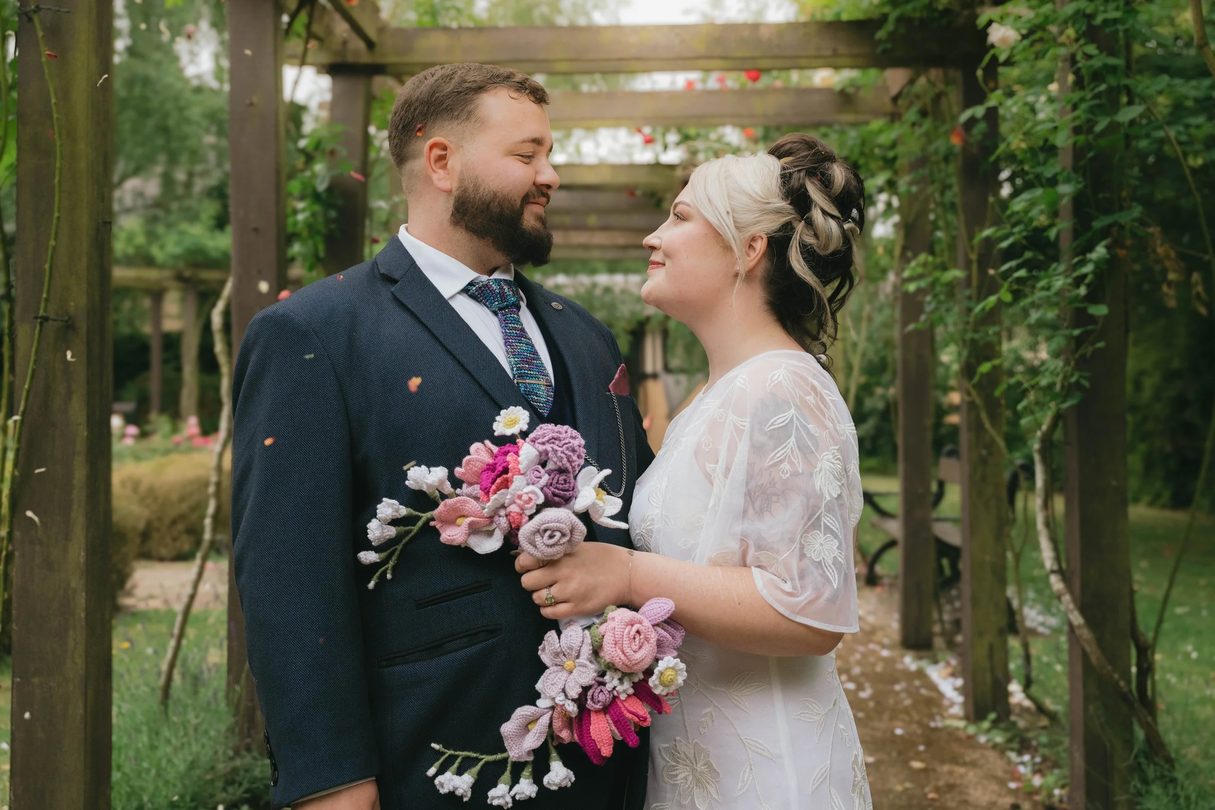 A couple in wedding attire standing close together in a garden, looking at each other, with the woman holding a bouquet of crocheted flowers.