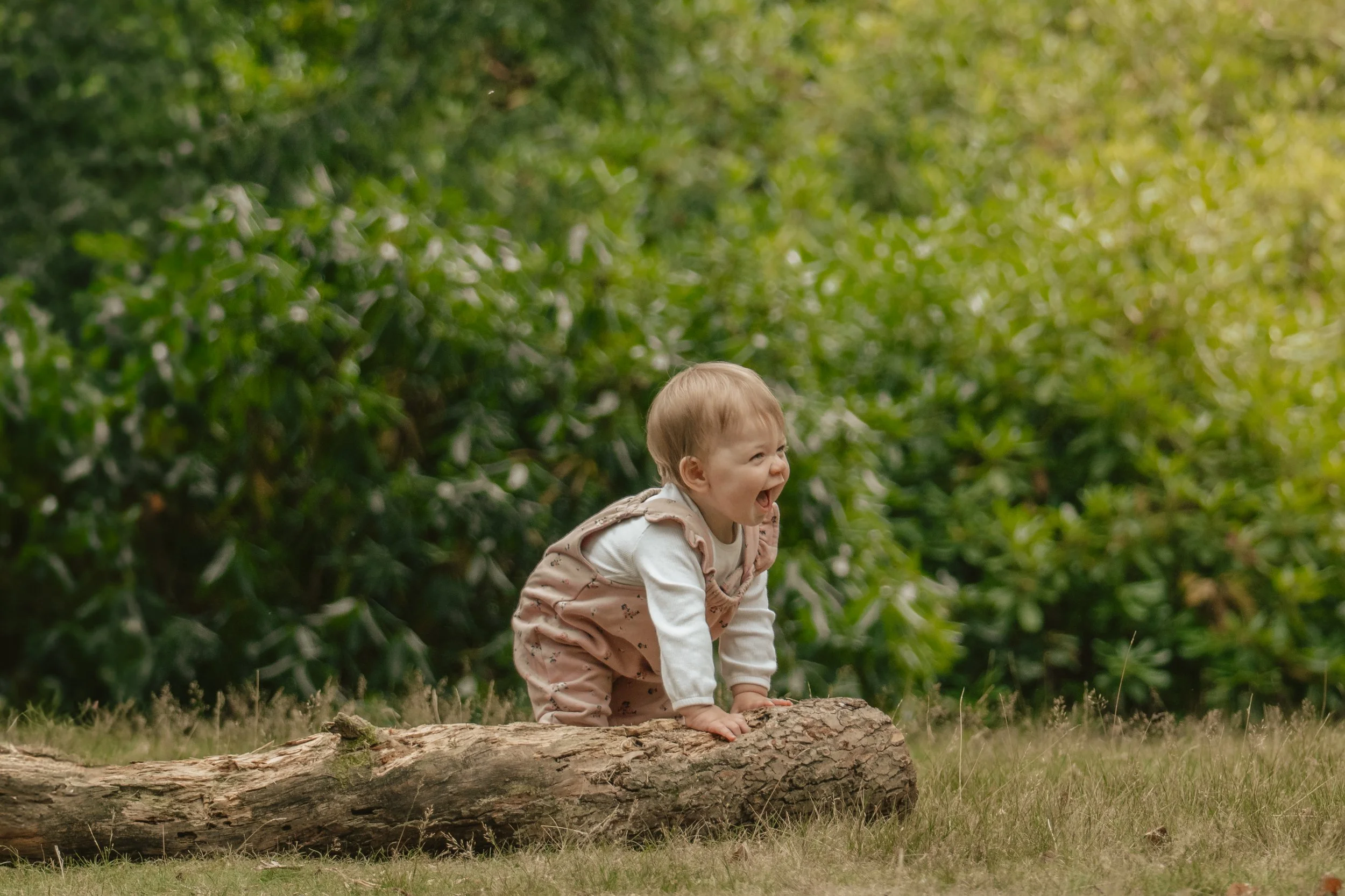 A young child in a white shirt and beige overalls with small patterns, crawling on a log in a lush green outdoor setting, with trees and foliage in the background, appearing happy and playful.