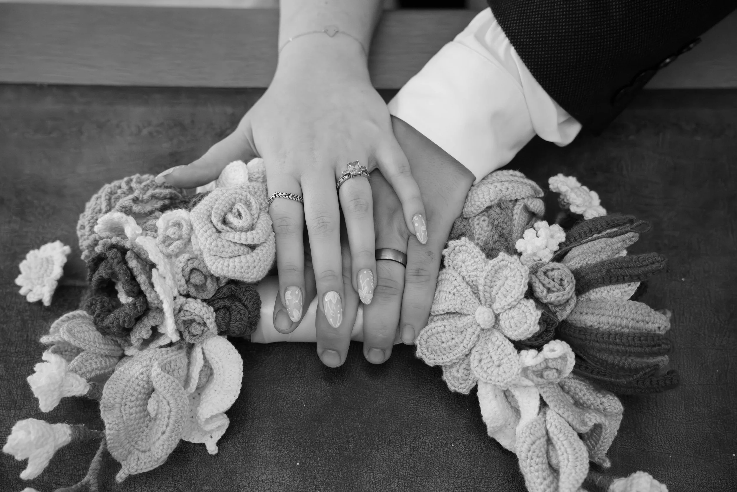 Two people are holding hands resting on a pile of knitted yarn scraps, with rings visible on their fingers. The person on top has long, decorated nails.
