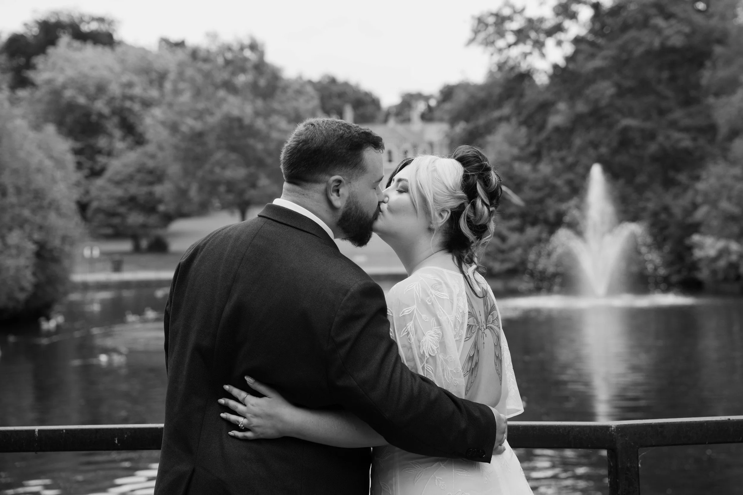 A couple wearing wedding attire sharing a kiss outdoors near a fountain with trees in the background.