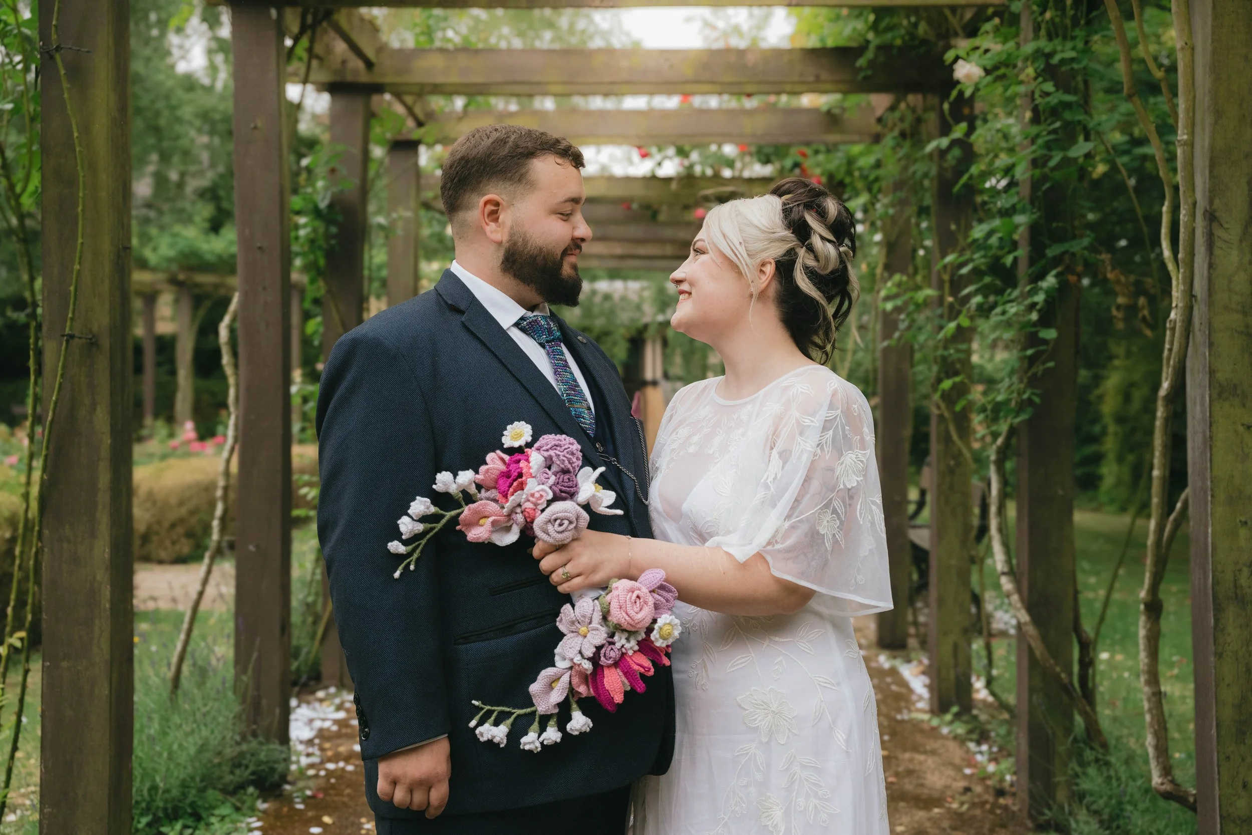 A newlywed couple smiling and looking into each other's eyes under a wooden garden pergola surrounded by greenery, with the bride holding a bouquet of pink and purple crochet flowers.