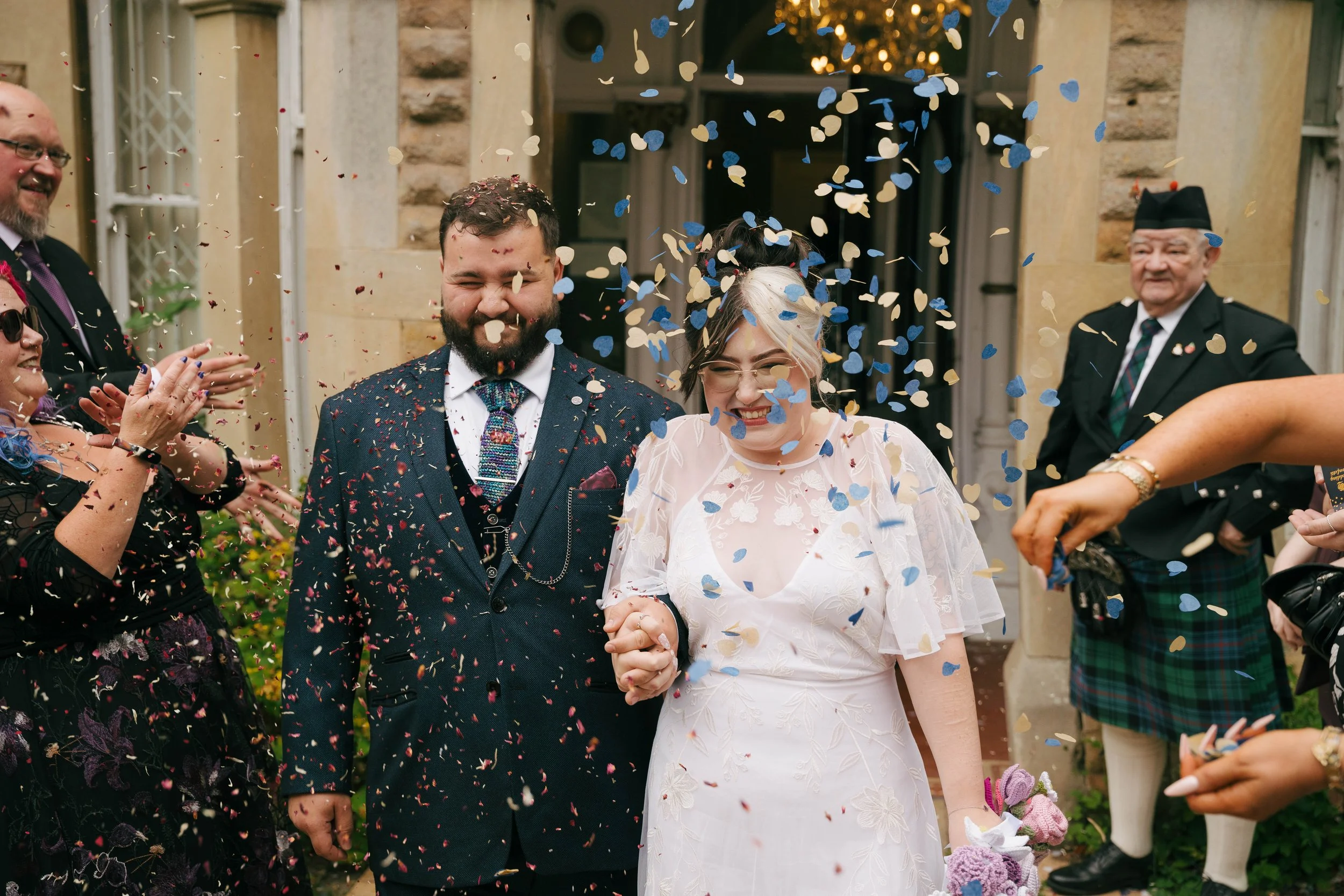 A newlywed couple holding hands, surrounded by confetti and guests celebrating outside a building.