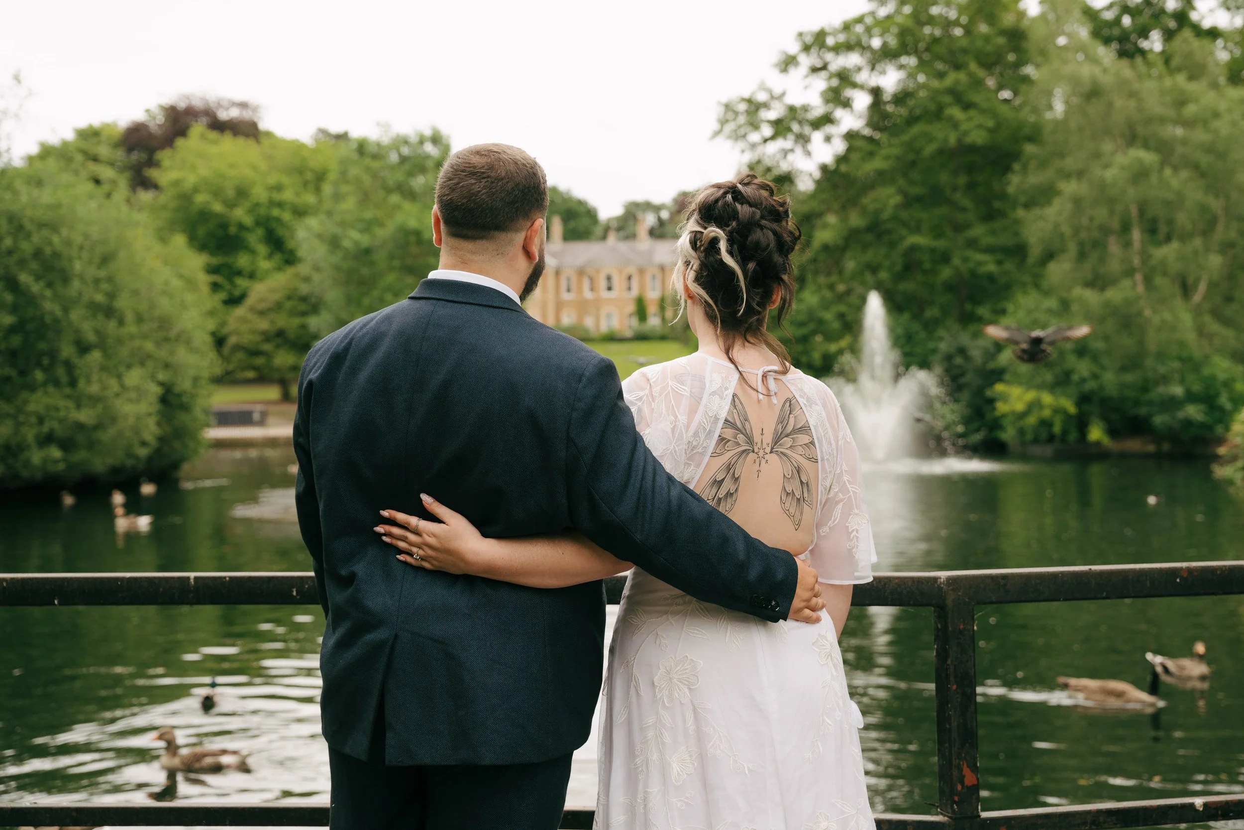 A couple dressed in wedding attire standing by a river in a park, with trees and a large historic building in the background.