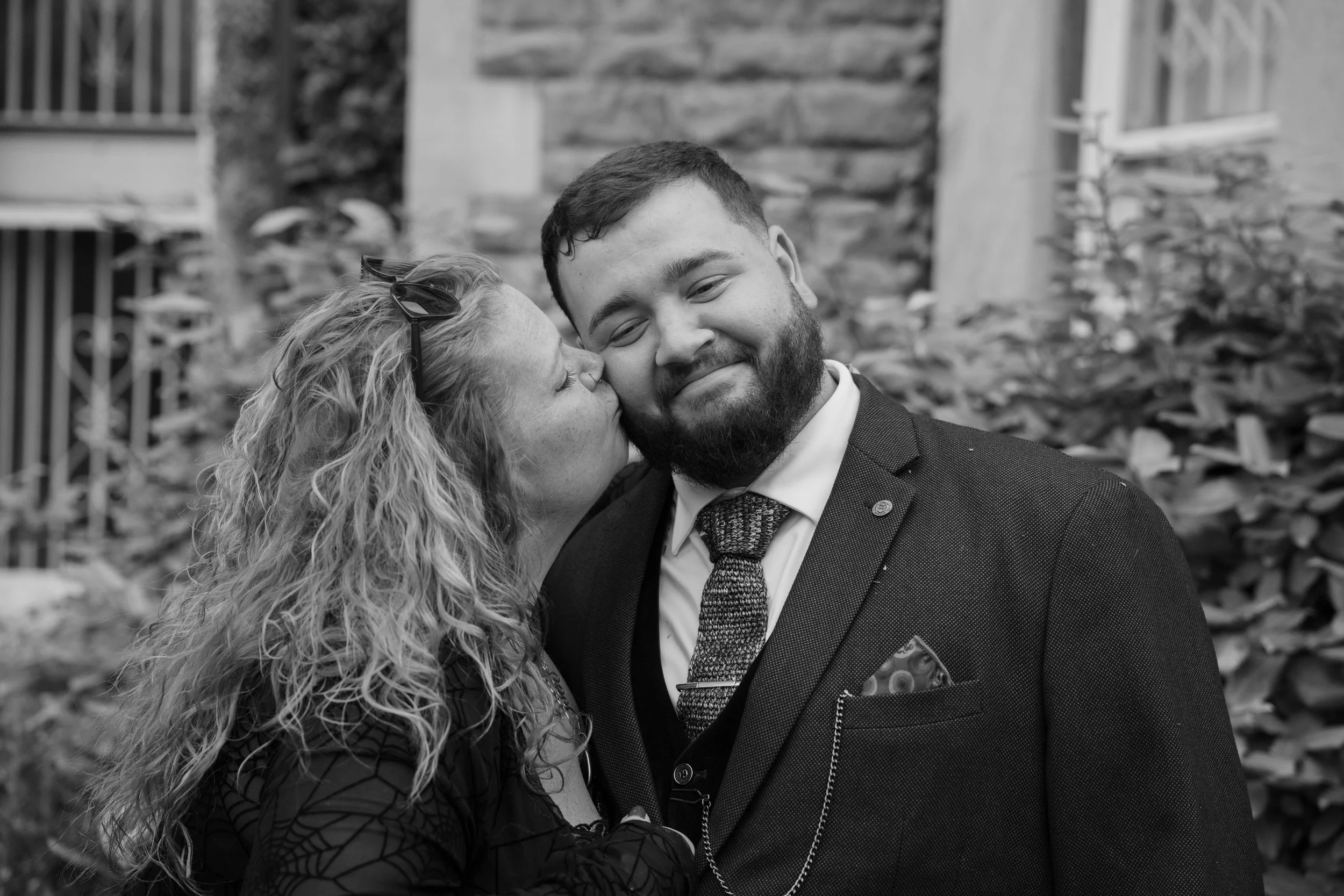 A black and white photo of a woman kissing a man on the cheek outdoors, with a background of stone and wooden garden structures.