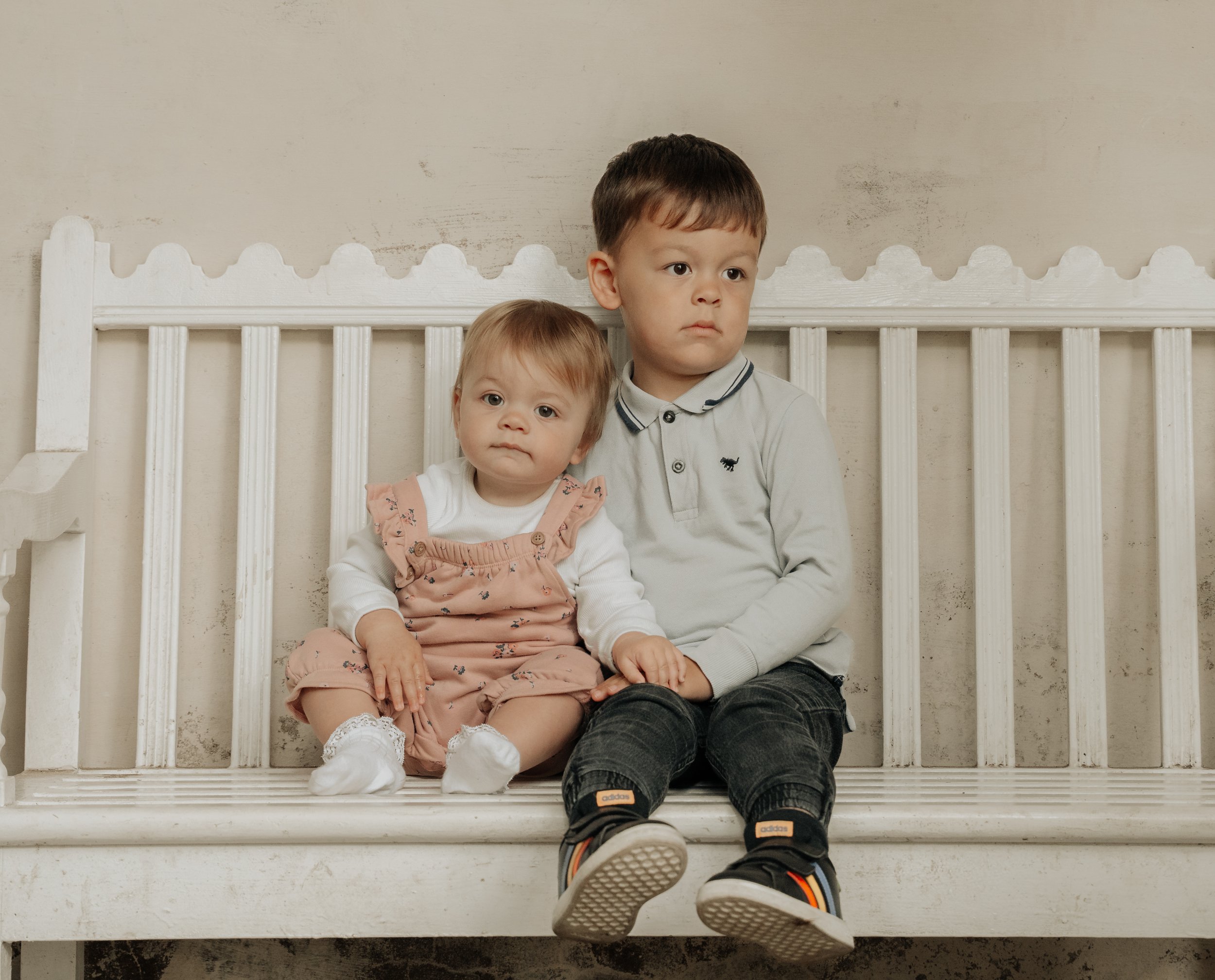 Two young children, a boy and a girl, sitting on a white wooden bench against a beige wall. The girl has light brown hair and wears a white shirt with pink overalls, while the boy has darker hair and is dressed in a light gray shirt with dark pants a