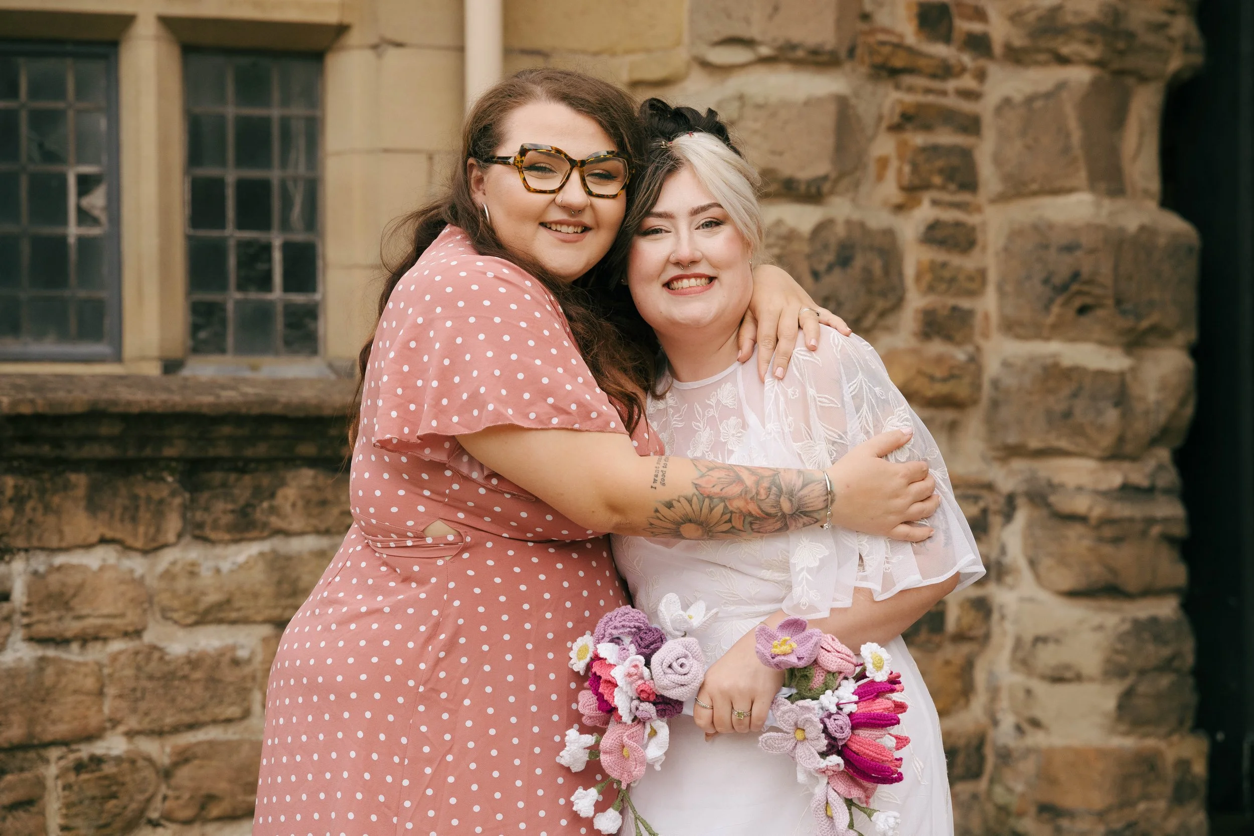 Two women smiling and hugging outside in front of a stone building. One woman has long dark hair, glasses, and tattoos on her arm; the other has short, two-tone black and white hair and is holding a bouquet of colorful crochet flowers.
