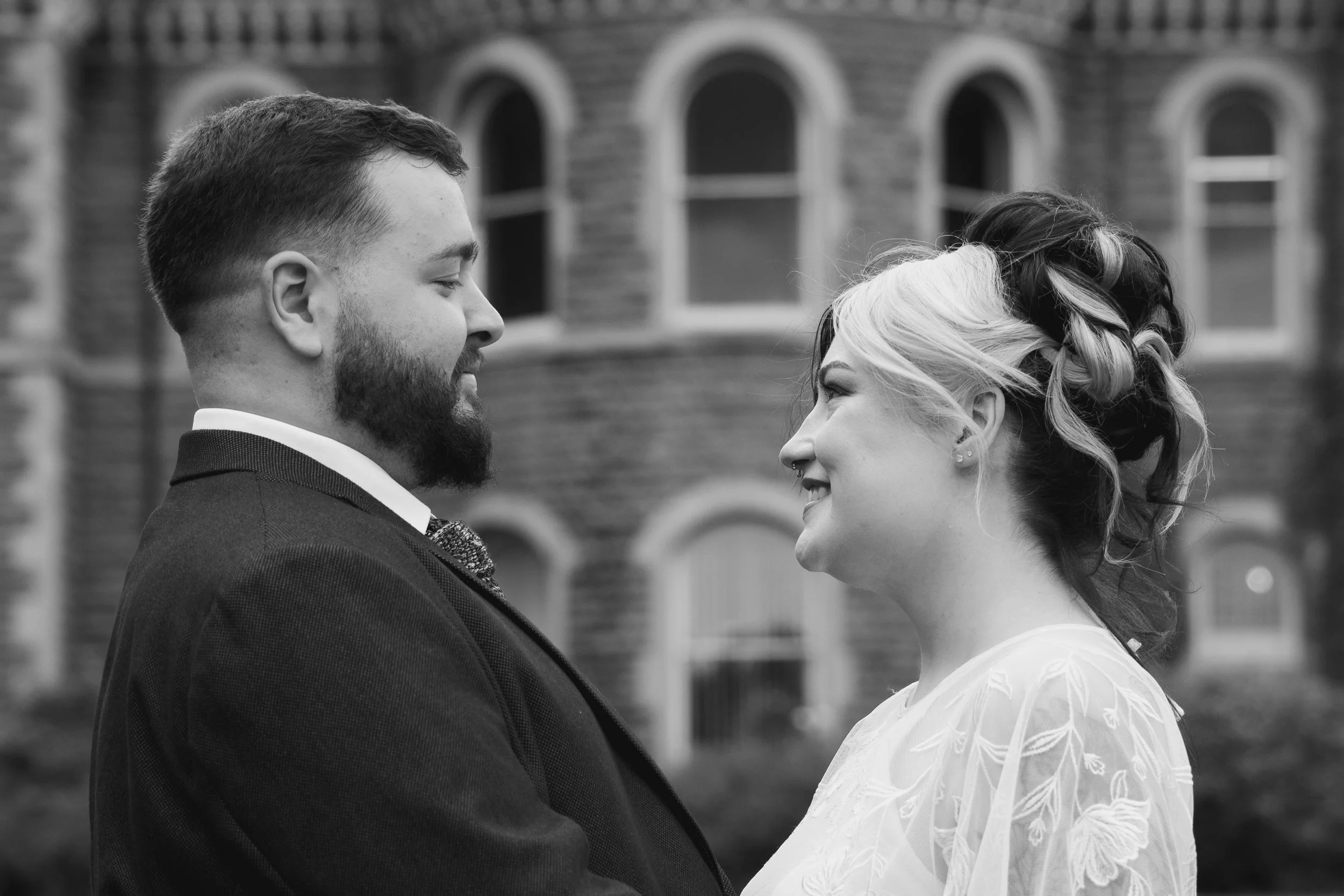 A black-and-white image of a man and woman standing face to face outdoors, smiling and looking into each other's eyes, with a brick building with arched windows in the background.