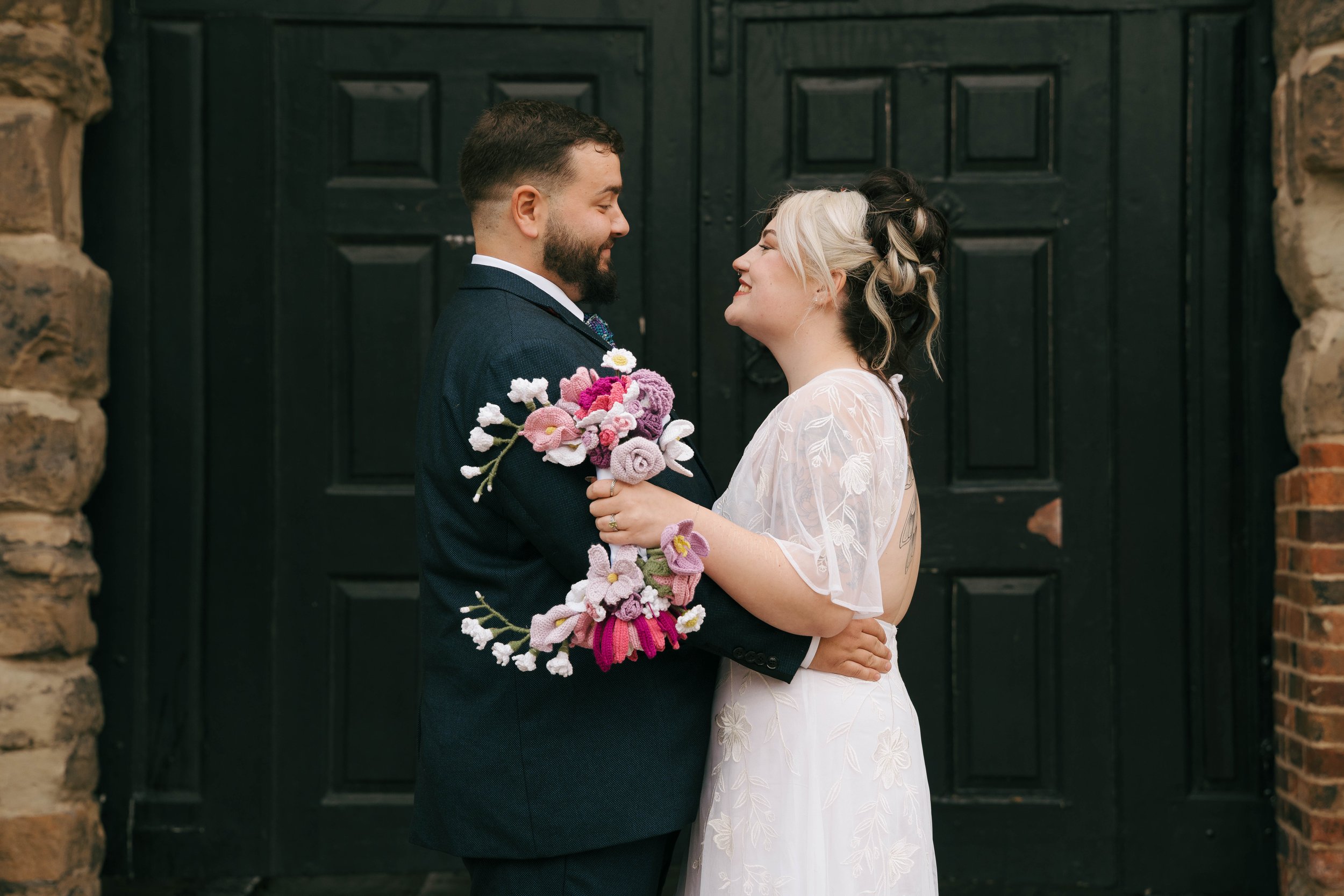 A bride and groom standing close, smiling at each other, in front of a black door with stone and brick walls on either side. The groom holds a bouquet of handmade felt flowers, and they are holding each other affectionately.