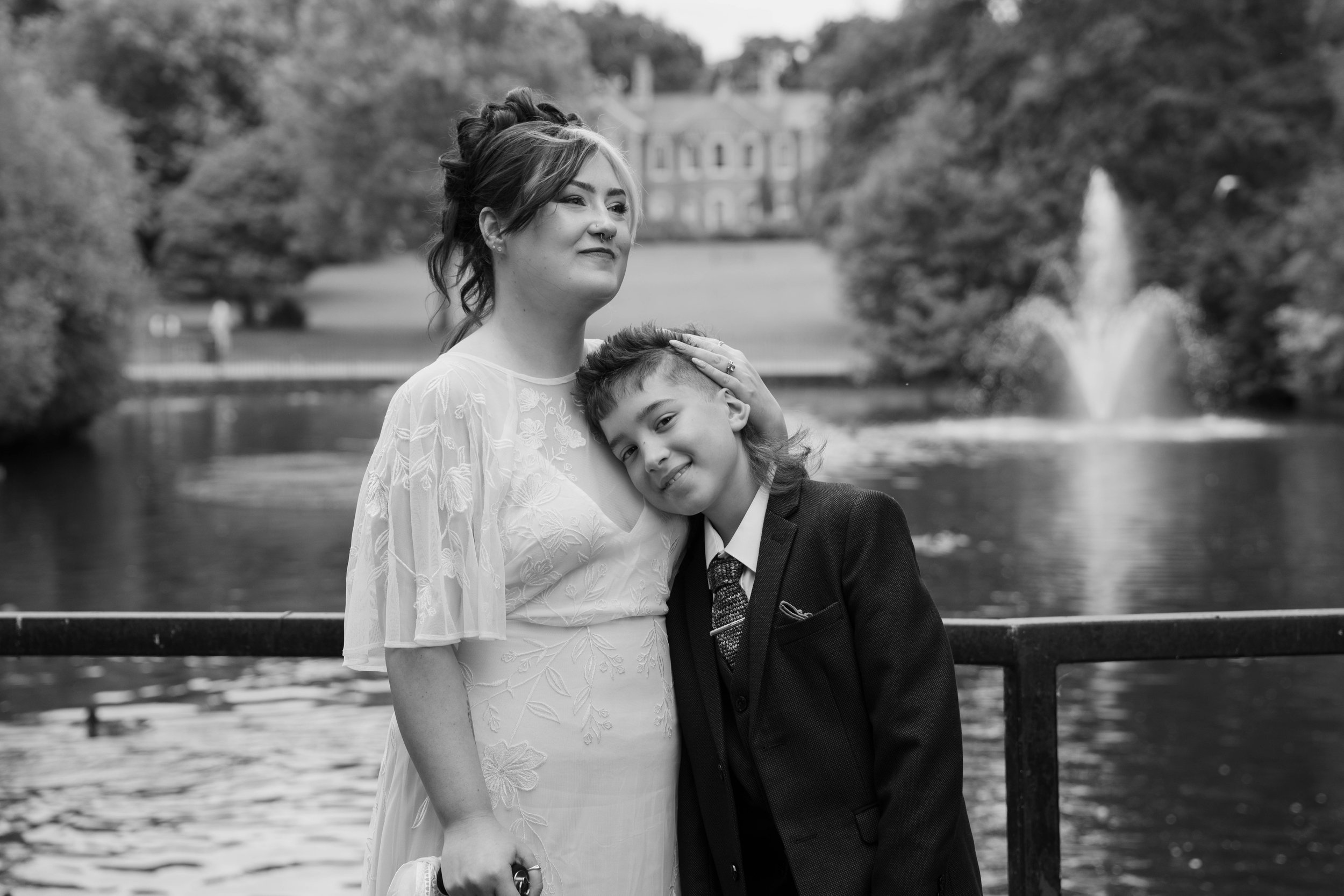 Black and white photo of a woman and a boy standing by a river, with the woman gently resting her hand on the boy's head, smiling softly, in a park with trees and a fountain in the background.
