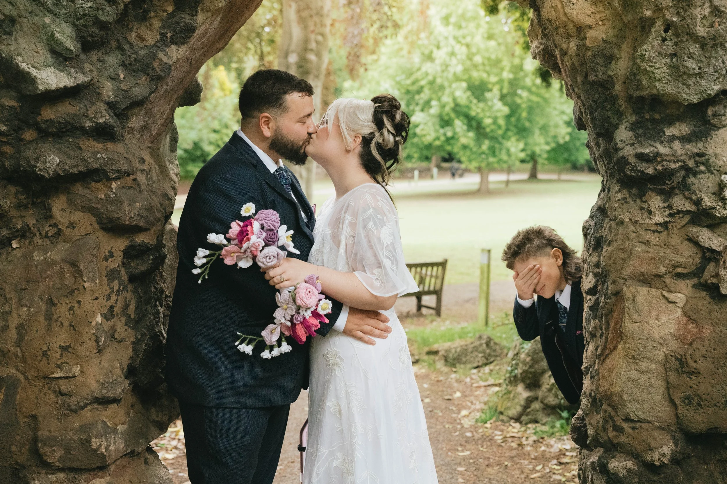 A bride and groom share a kiss in an outdoor park, framed by a large rock formation with greenery and trees in the background, while a young boy covers his face nearby.