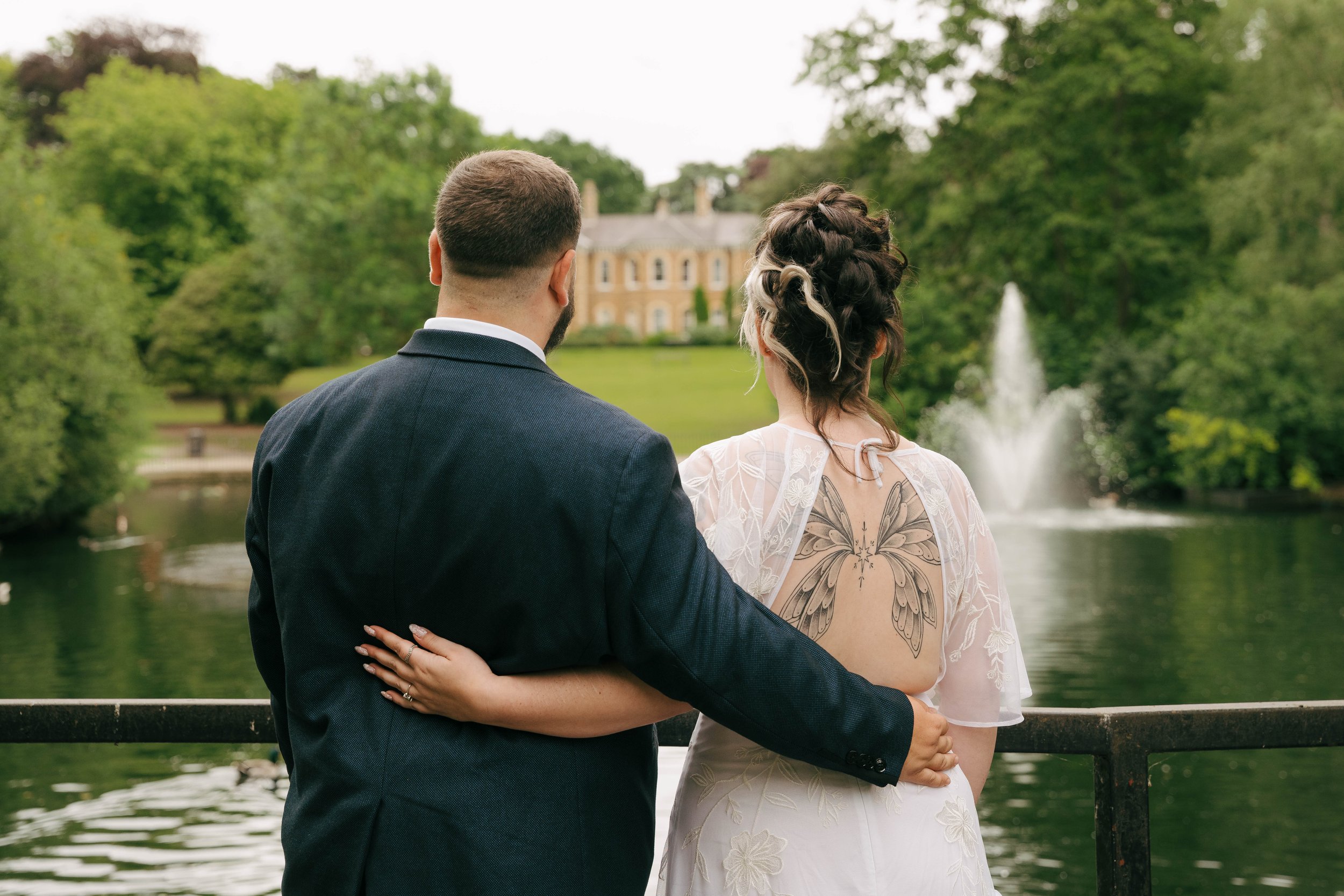 A couple, seen from behind, standing close together by a lake with a fountain, surrounded by trees, with a large house in the background.