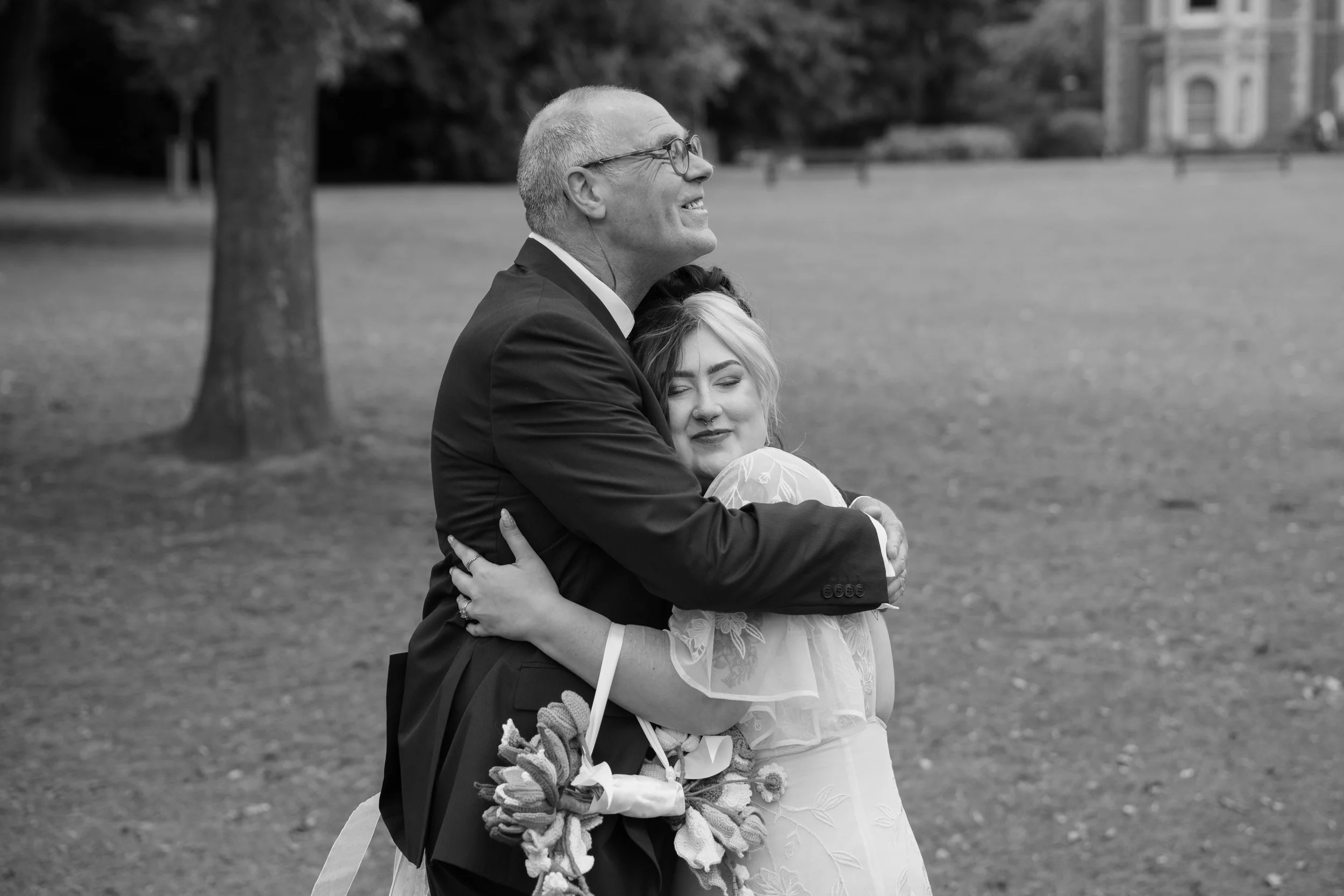 A man in glasses and a suit embraces a woman in a light-colored dress and gloves in a park with trees and a building in the background.