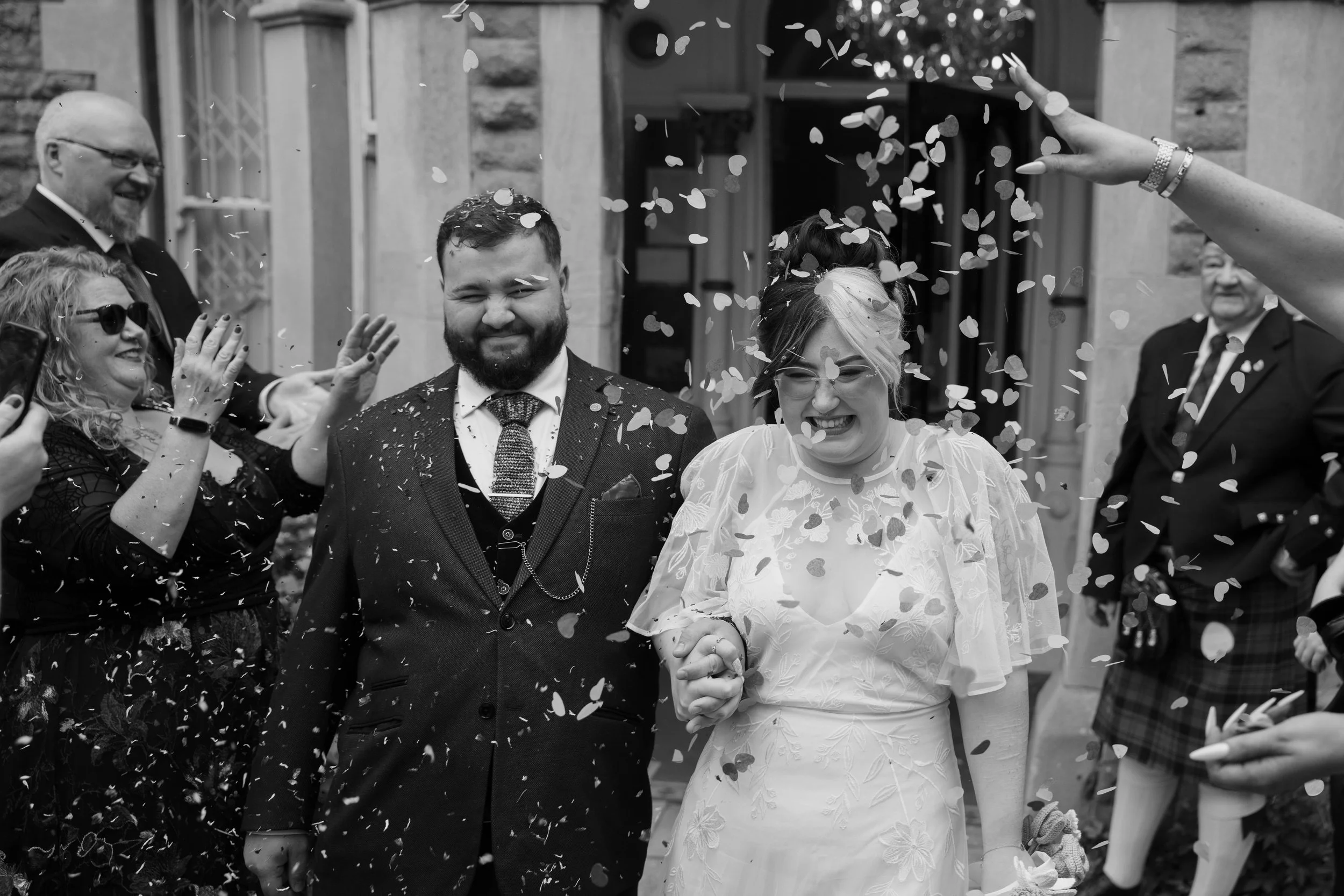 A black and white photo of a group of people celebrating a wedding. The bride and groom are holding hands and smiling as confetti is thrown around them. The wedding is outside a building with stone walls.