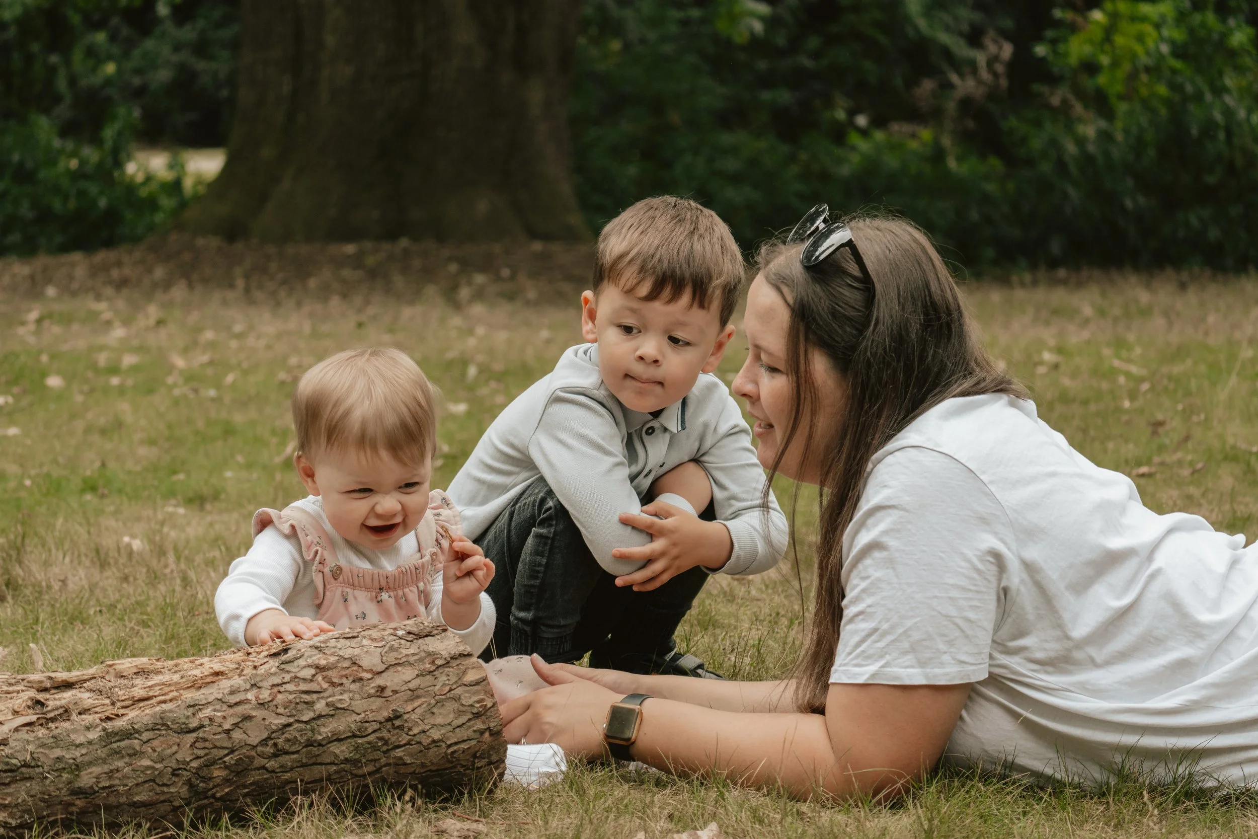 A woman with sunglasses on her head is lying on the grass with two young children, one of whom is sitting on her back and another is reaching for a log in a wooded park.