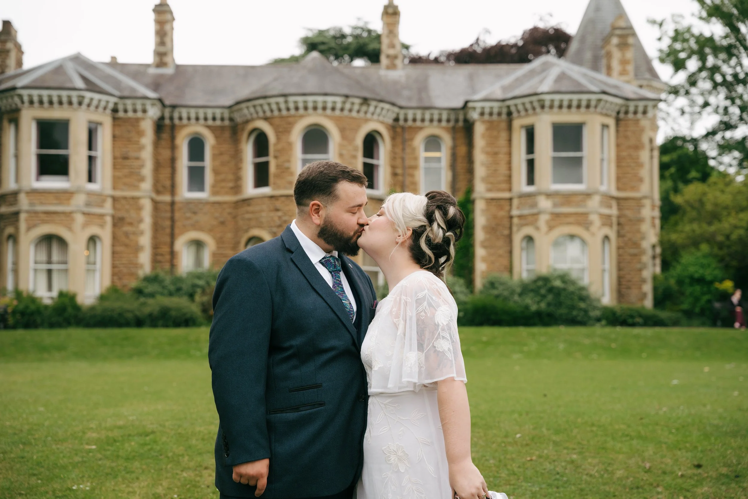 A couple in wedding attire sharing a kiss outdoors in front of a large historic brick building with multiple windows and towers.