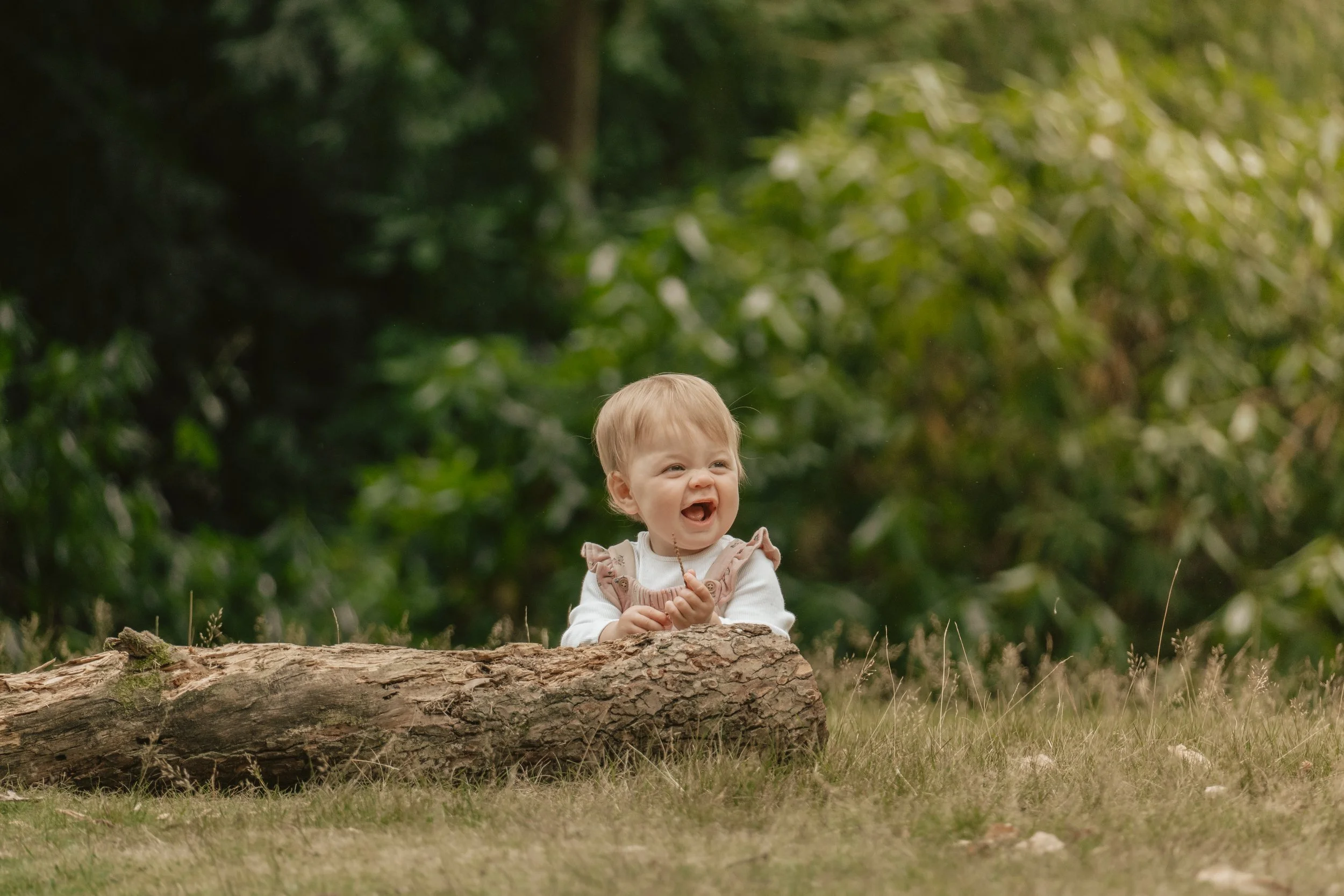 A young girl with blonde hair smiling and laughing while sitting behind a large fallen tree trunk in a forested area surrounded by green foliage.