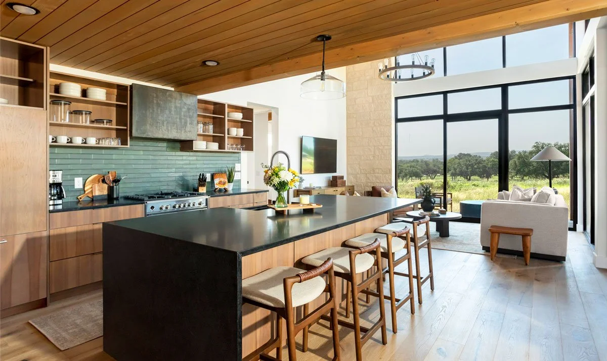 Open-concept kitchen and living area with large windows, wooden ceiling, black island countertop, and modern furnishings overlooking a green landscape in Hye Springs Ranch in Fredericksburg TX.