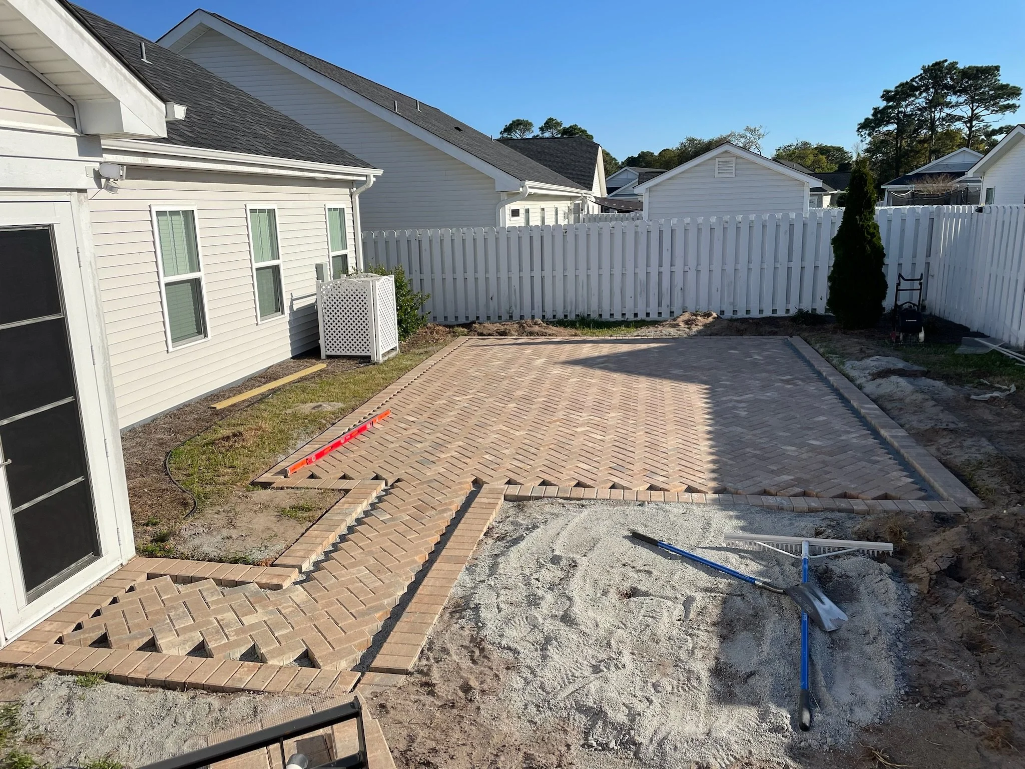 Backyard patio construction with brick paving in progress, including tools and partially laid bricks, surrounded by white fence and house exterior.