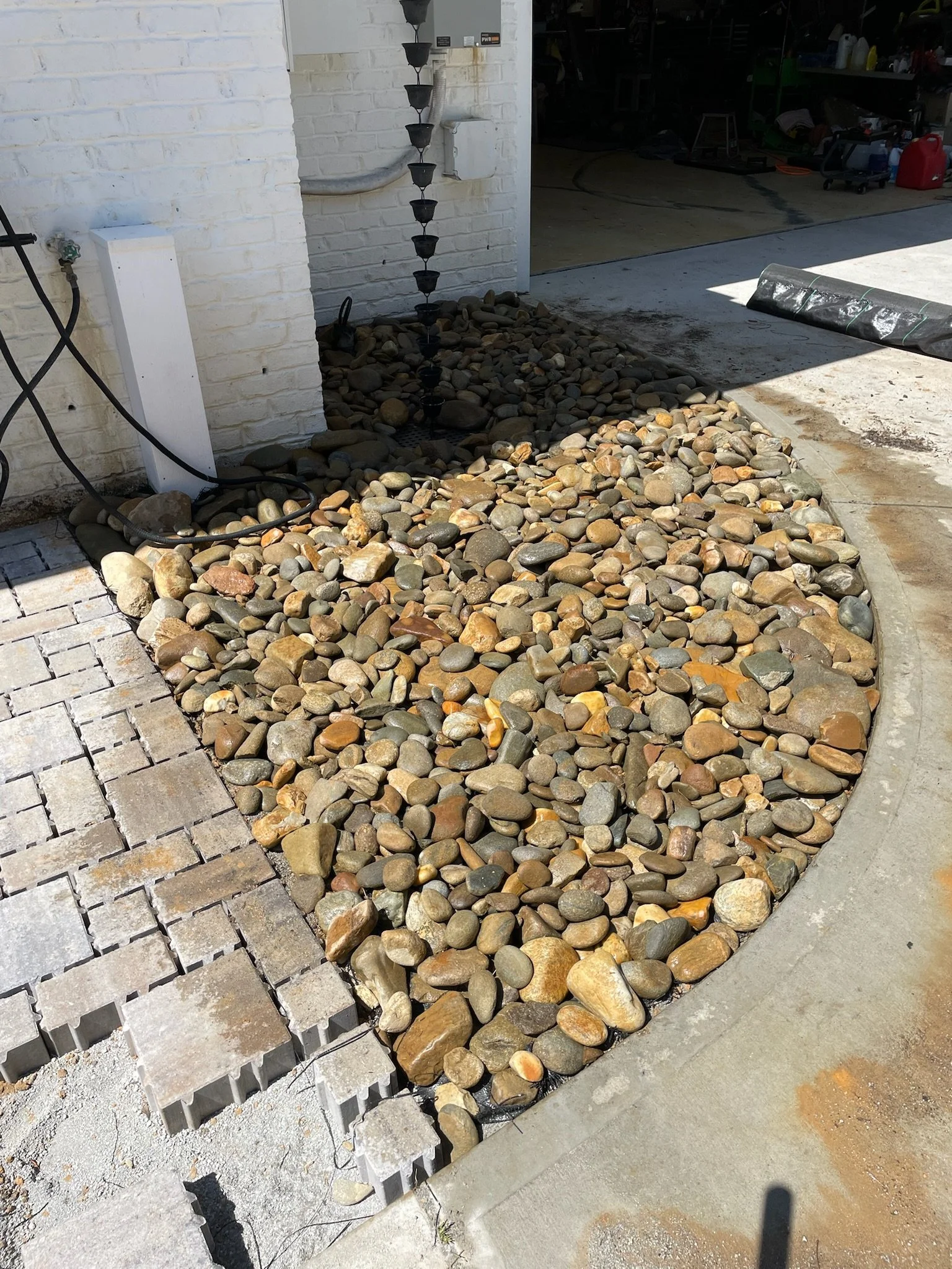 Landscape area with decorative stones, concrete edging, and brick pavers in front of an exterior wall with a black rain chain.