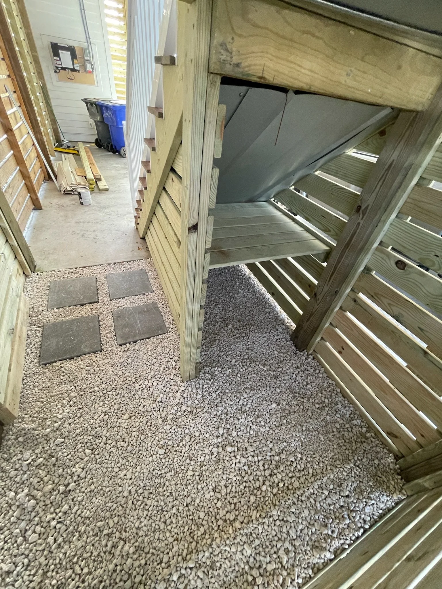 Backyard area with wooden stairs and slat fence, gravel floor, utility bin, and tools on concrete surface.