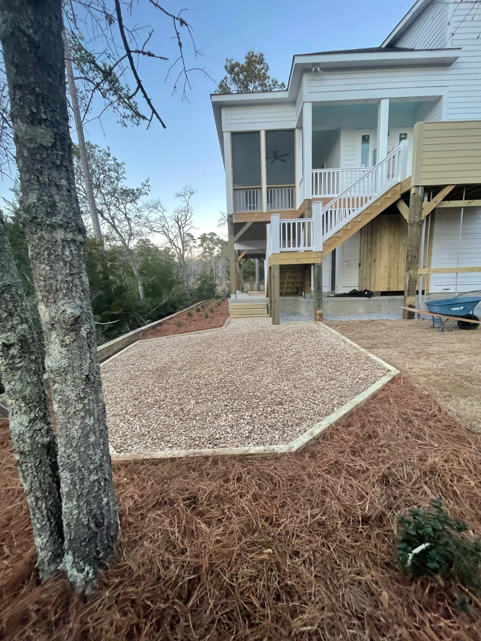 Elevated modern white house with wooden stairs, overlooking a landscaped yard with gravel and mulch, surrounded by trees.