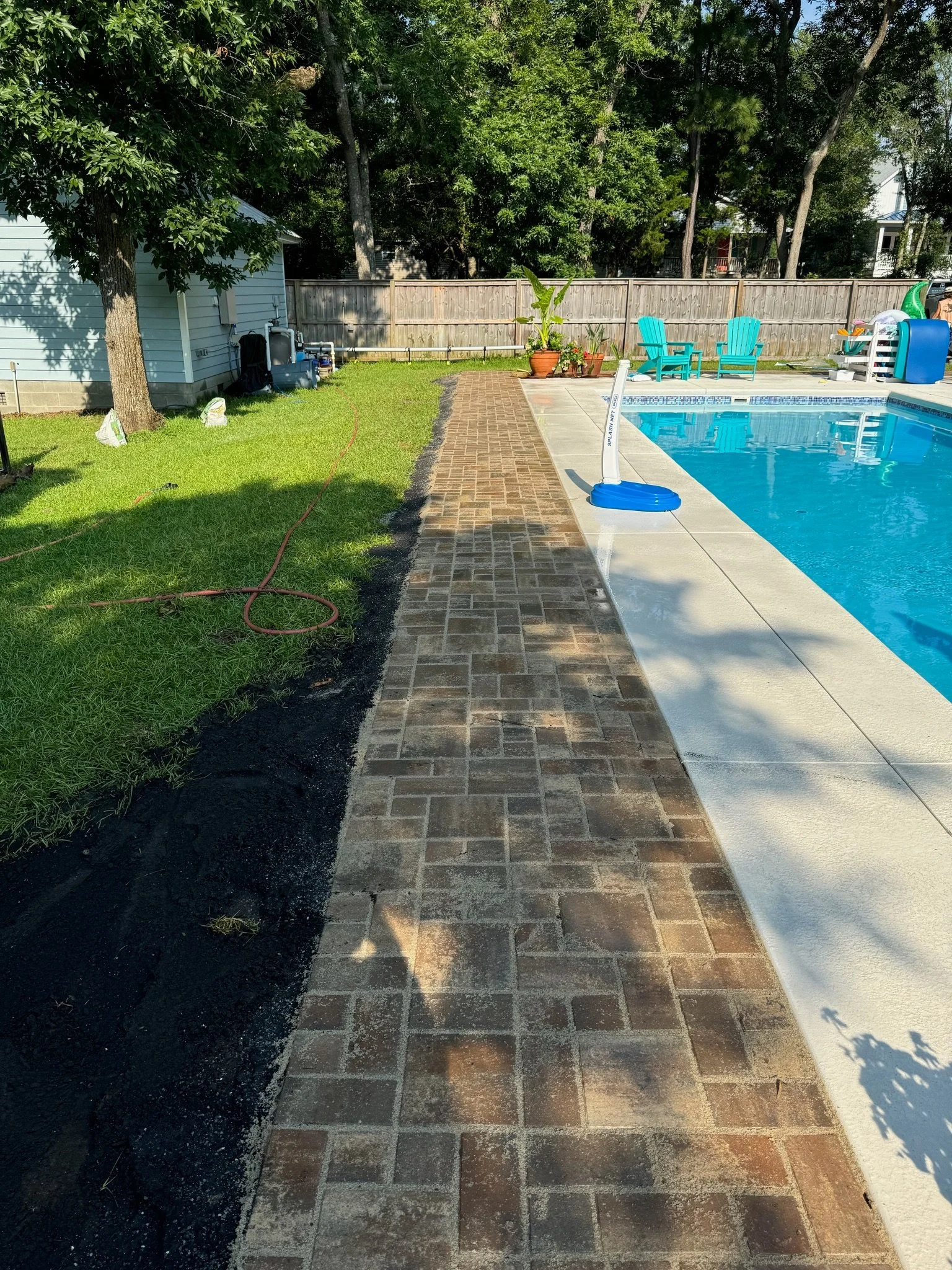 Brick pathway next to a swimming pool, with a grassy yard on the left, trees in the background, and blue chairs near the pool.