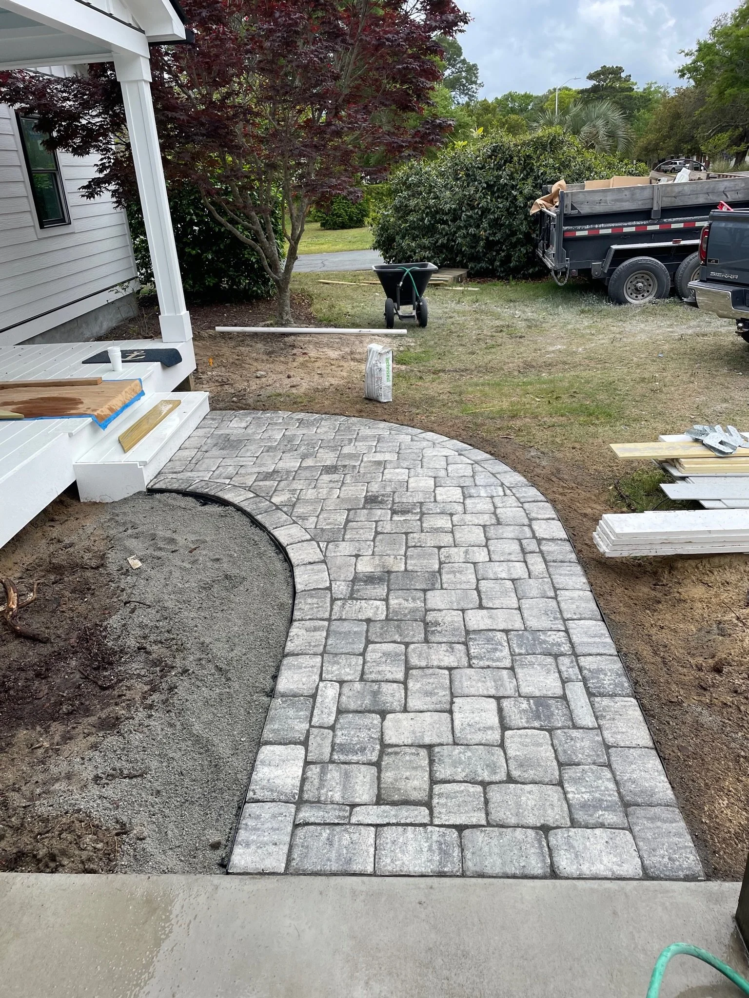 A partially completed garden pathway made of stone pavers curves from a porch. A wheelbarrow and a pile of construction materials are visible near a truck in the background, with trees and bushes surrounding the area.