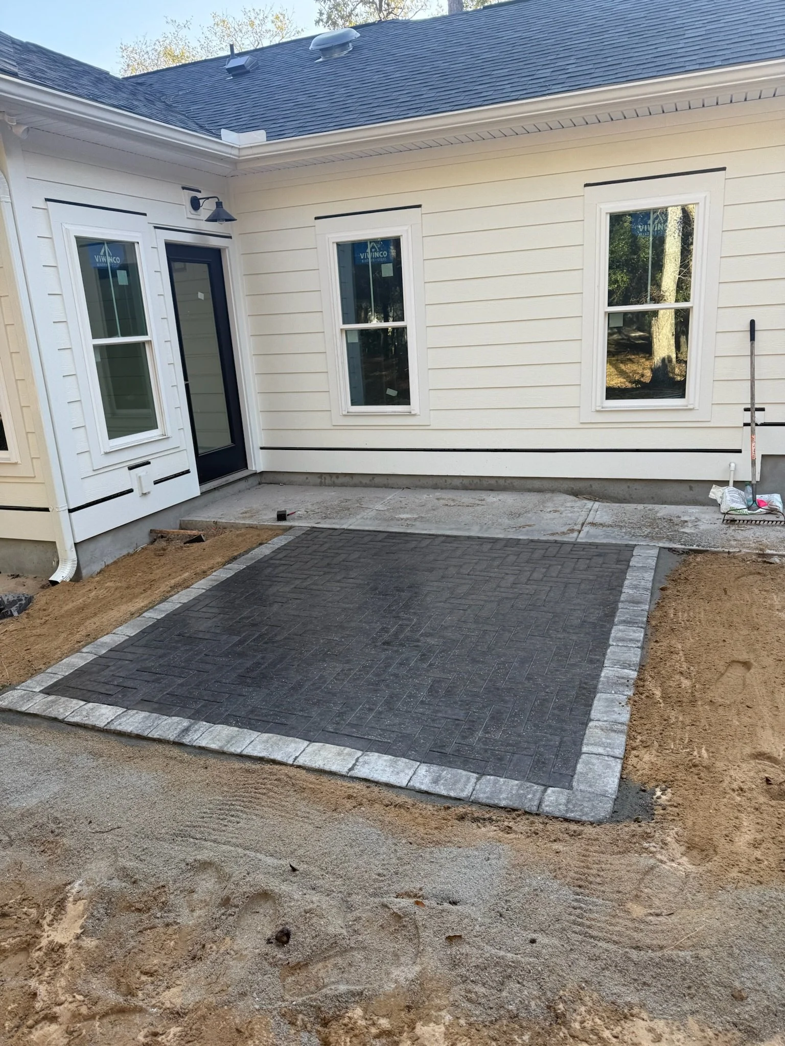 Newly installed concrete patio with decorative brick pattern in a backyard, adjacent to a white house with multiple windows.