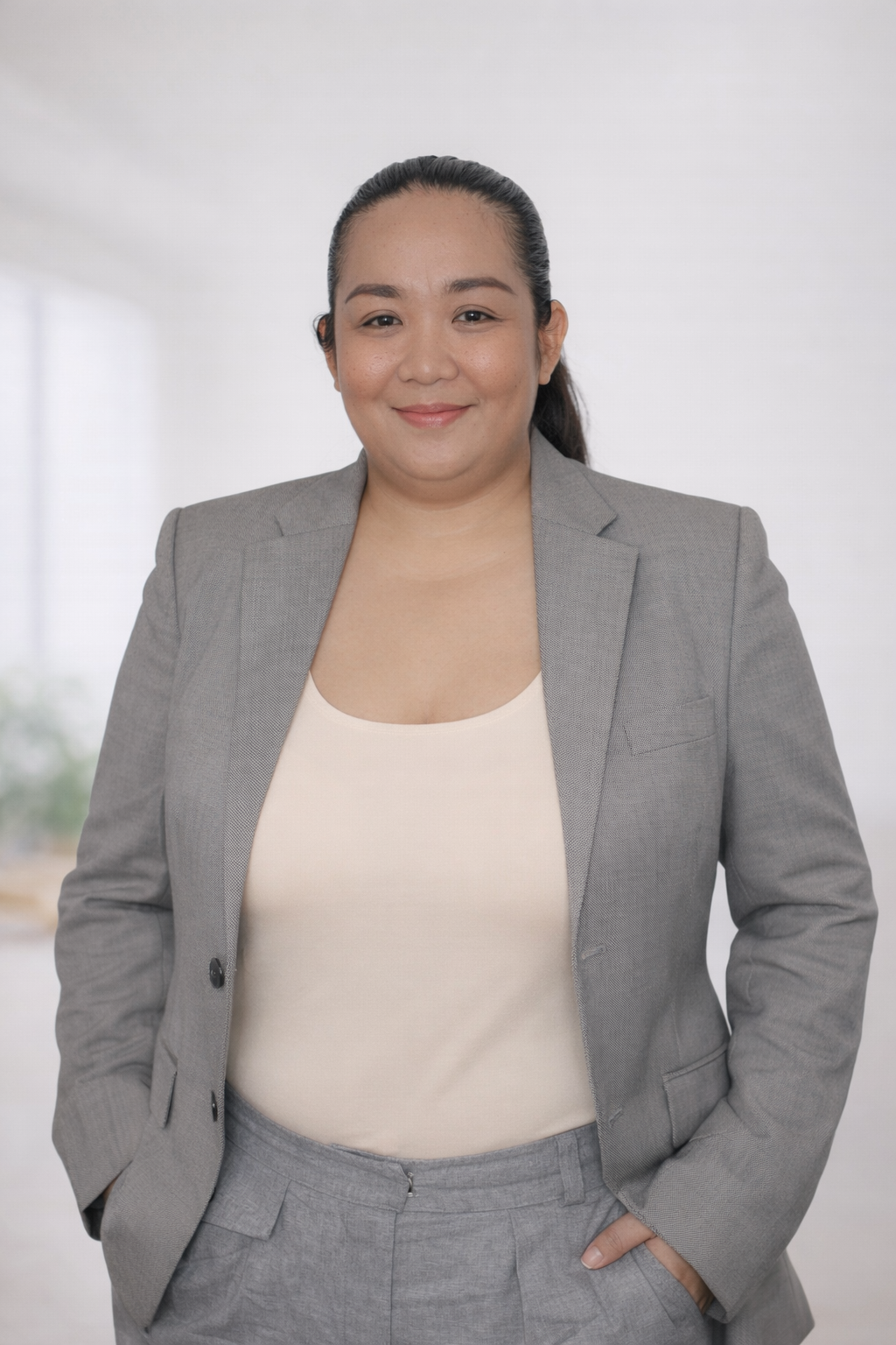 A woman in a light gray business suit and cream top standing with hands in pockets, smiling in an office setting.