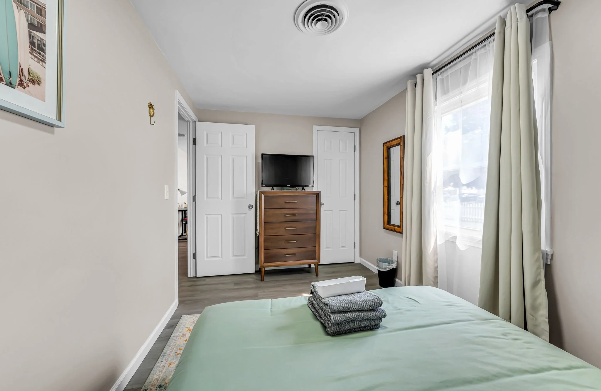 A bedroom with light-colored walls and a wooden dresser with a TV on top. There is a window with white curtains on the right side, and a door and closet doors in the background. Part of a bed with folded gray towels is visible in the foreground.
