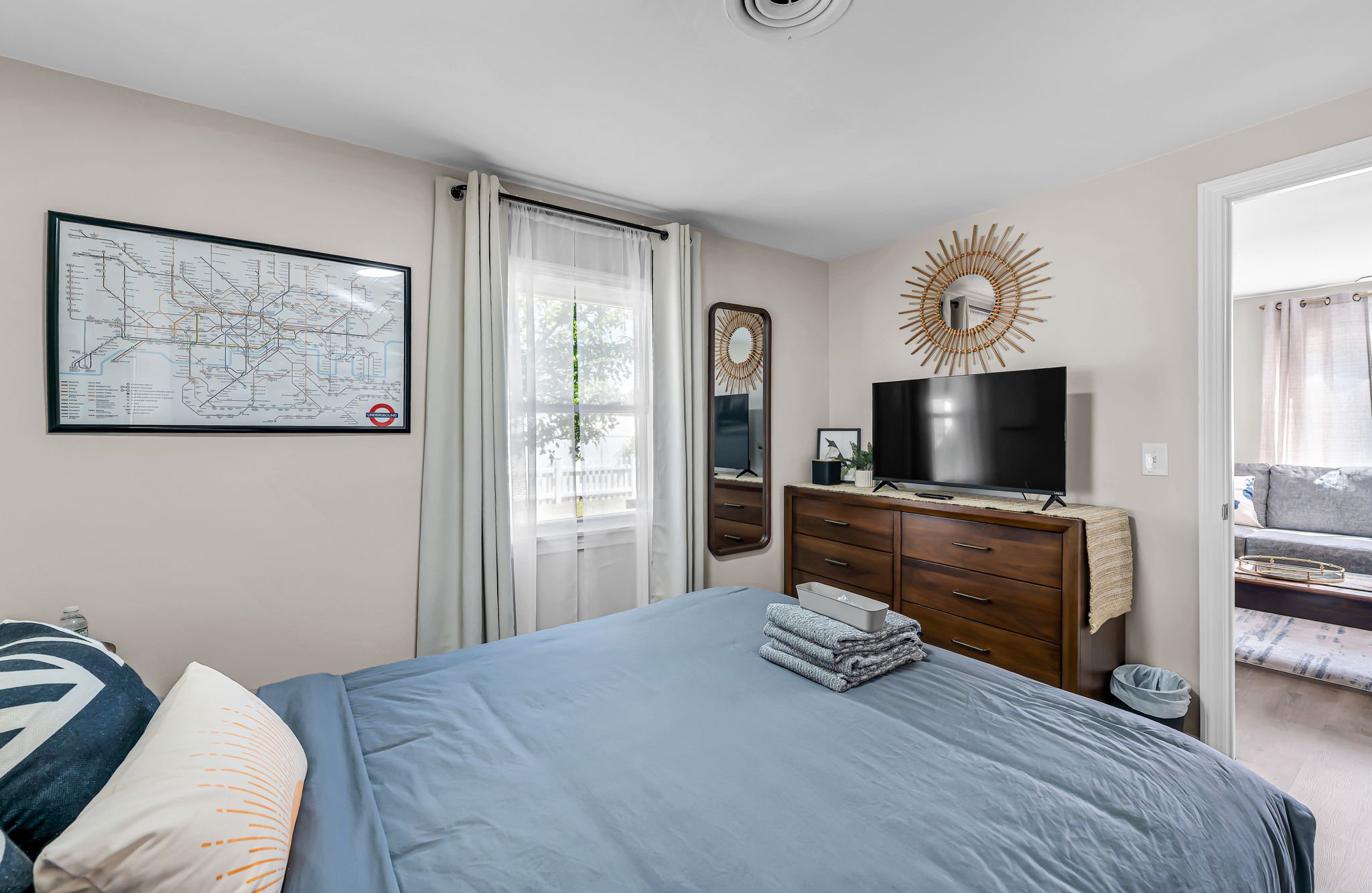 A bedroom with a bed, pillows, and neatly folded towels on top of the bed. The room has a window with white curtains, a dresser with a flat-screen TV, a decorative round mirror, and wall art resembling a subway map. There is a small mirror on the wal