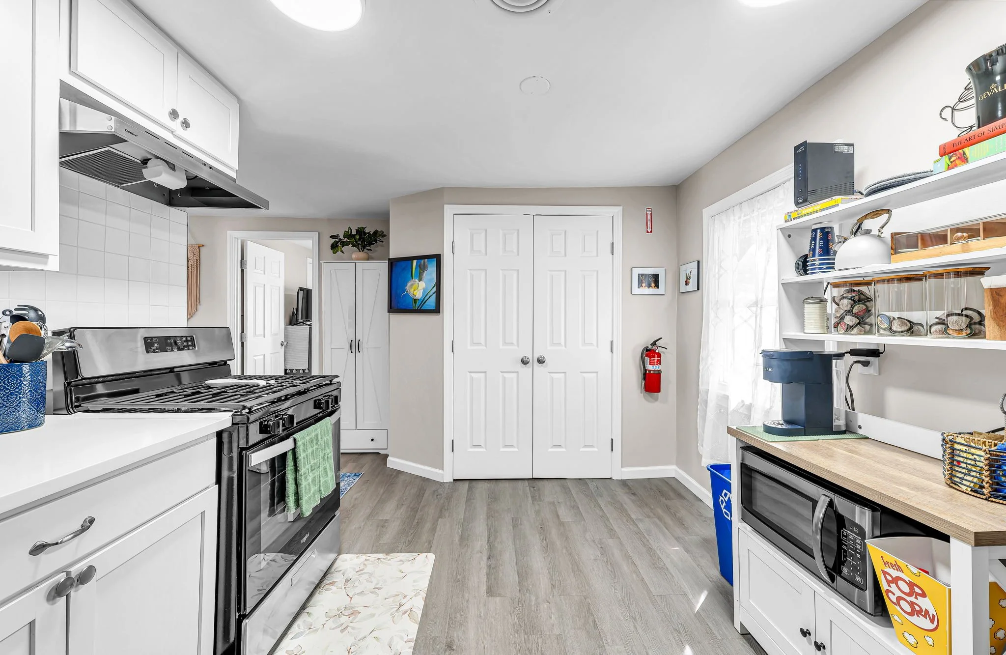 A bright, modern kitchen with white cabinets, a black stove, and wood flooring. Shelves with dishes, a coffee machine, and books are on the right wall, and a small TV is mounted on the wall near a set of closed white double doors. Kitchen accessories