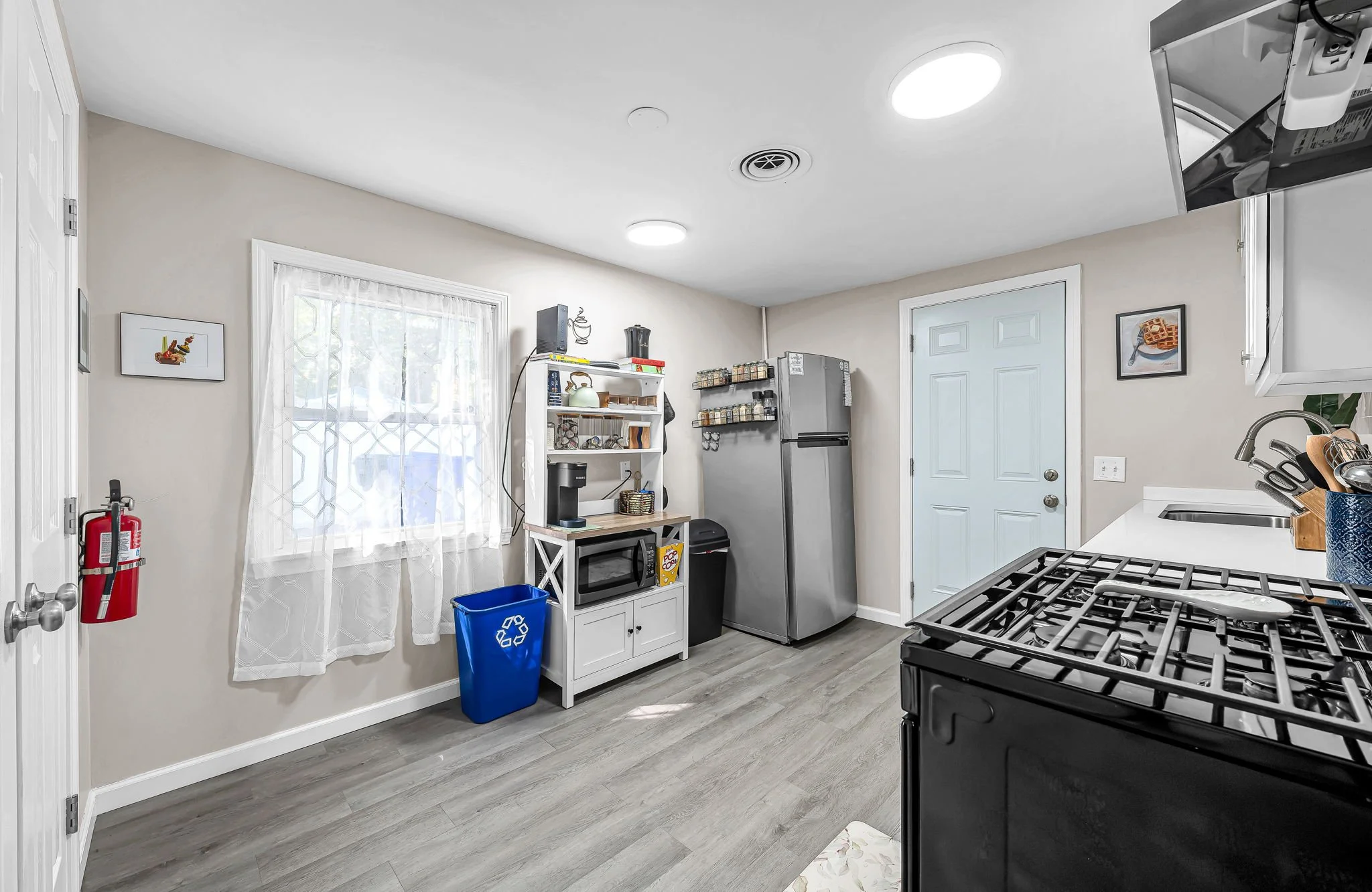 A kitchen with a window, a white bookshelf, a fridge, a microwave, a stove, and a double sink. The walls are beige, and the flooring is gray wood.